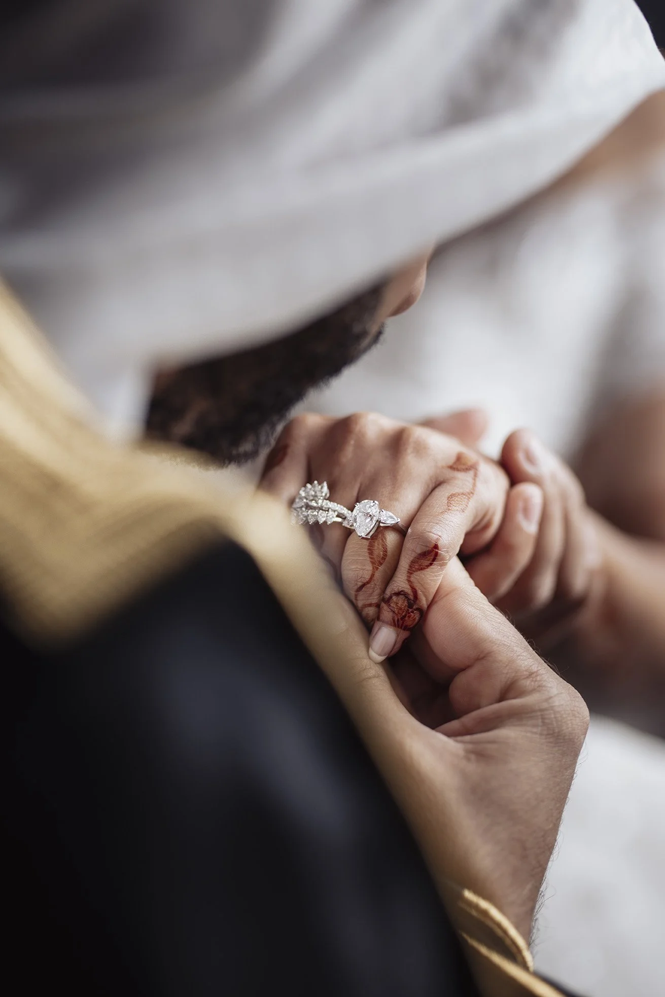A close-up of a bride and groom holding hands, with the groom wearing a diamond engagement ring. The bride's hand has henna designs.