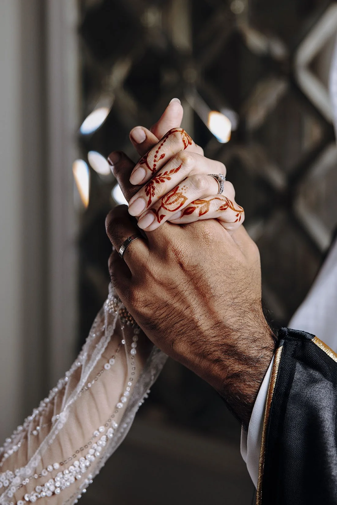 Close-up of a couple holding hands, showing detailed henna designs on their fingers. The woman’s hand, adorned with a wedding ring and embellished sleeve, rests on top of the man’s hand, which is large, with a wedding band. The background features dark, geometric shapes with soft lighting.