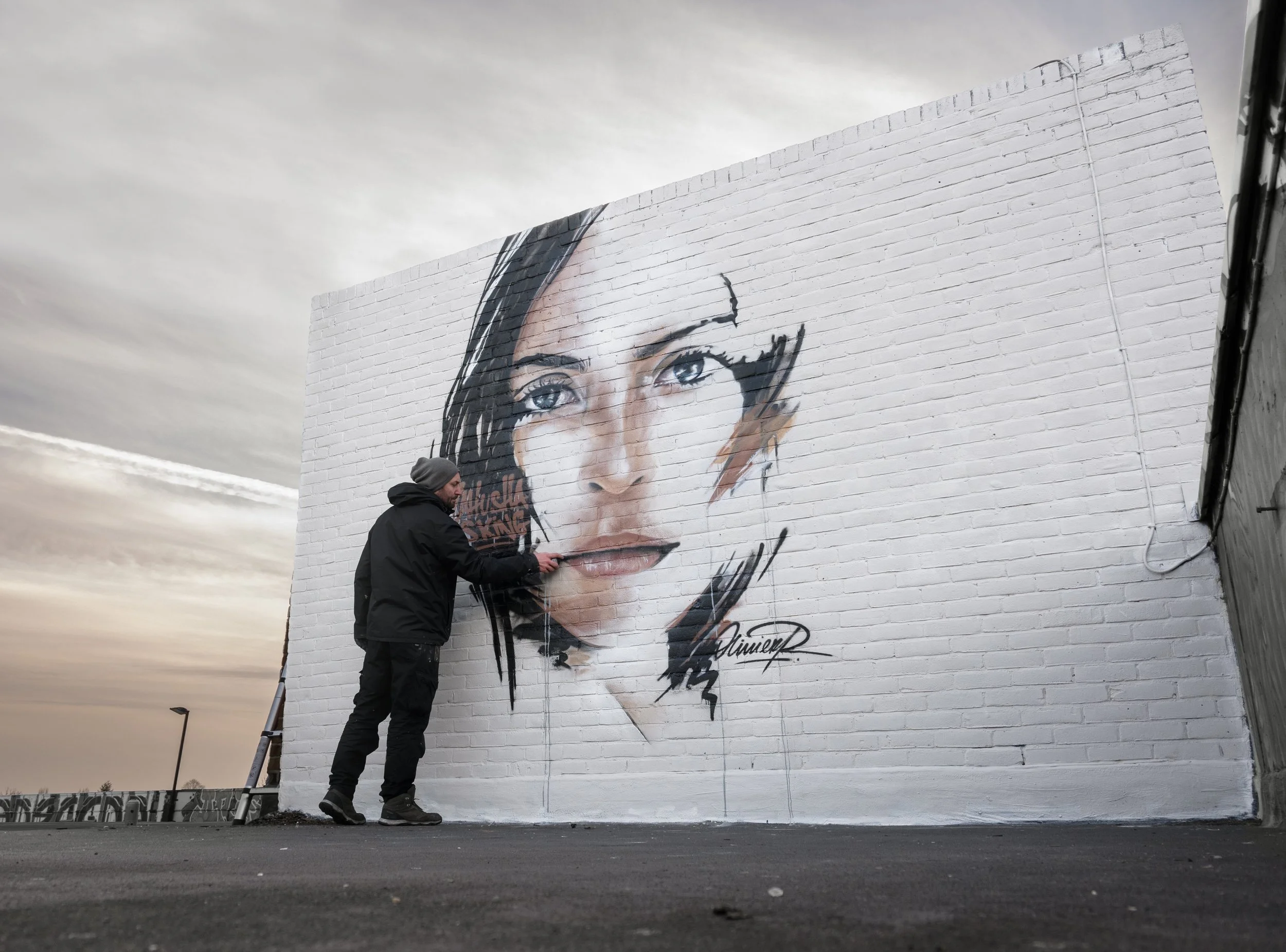 Street artist painting a large portrait of a woman on a white brick wall during sunset
