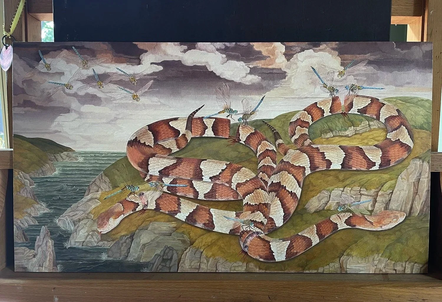 A painting of a coiled rattlesnake on a rocky hillside near water, surrounded by flying dragonflies under a cloudy sky.