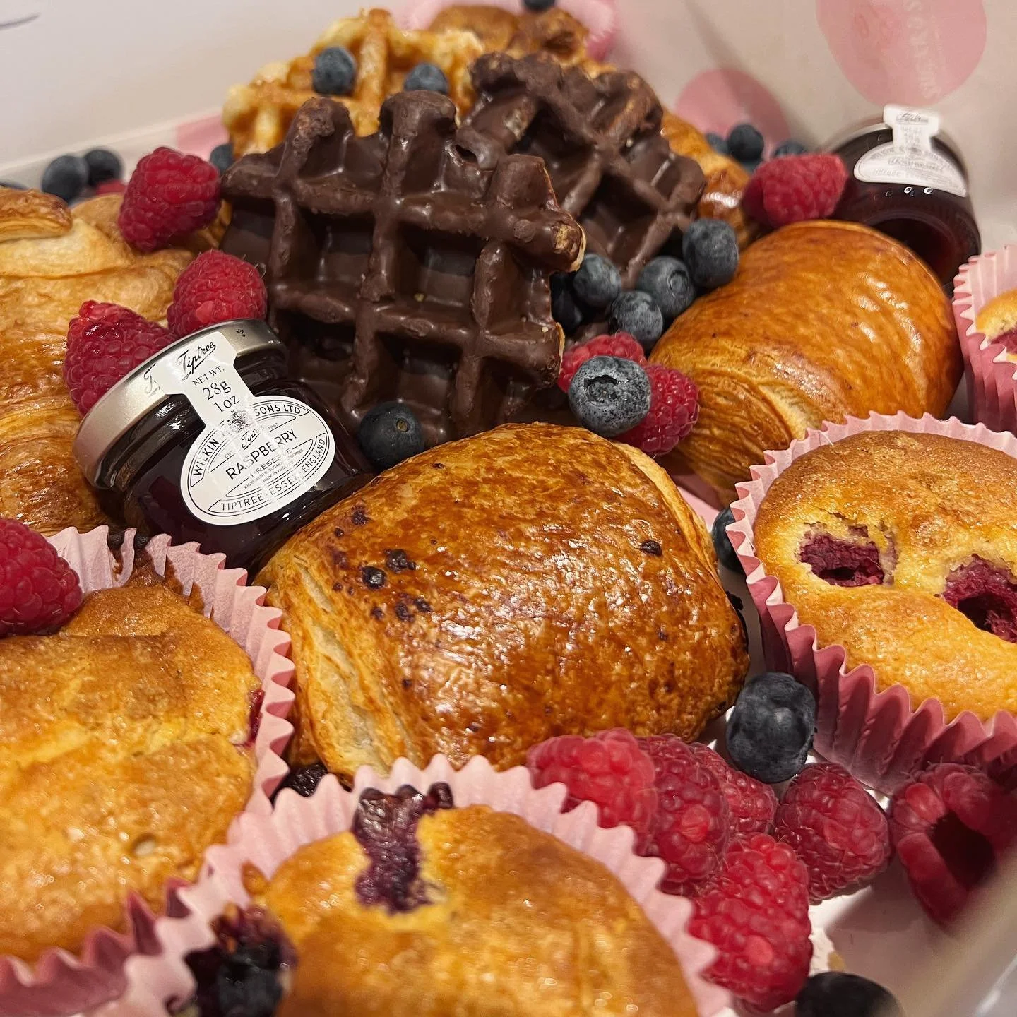A variety of pastries including croissants, muffins, and chocolate-covered waffles surrounded by fresh raspberries and blueberries. A small jar of raspberry jam is also visible.