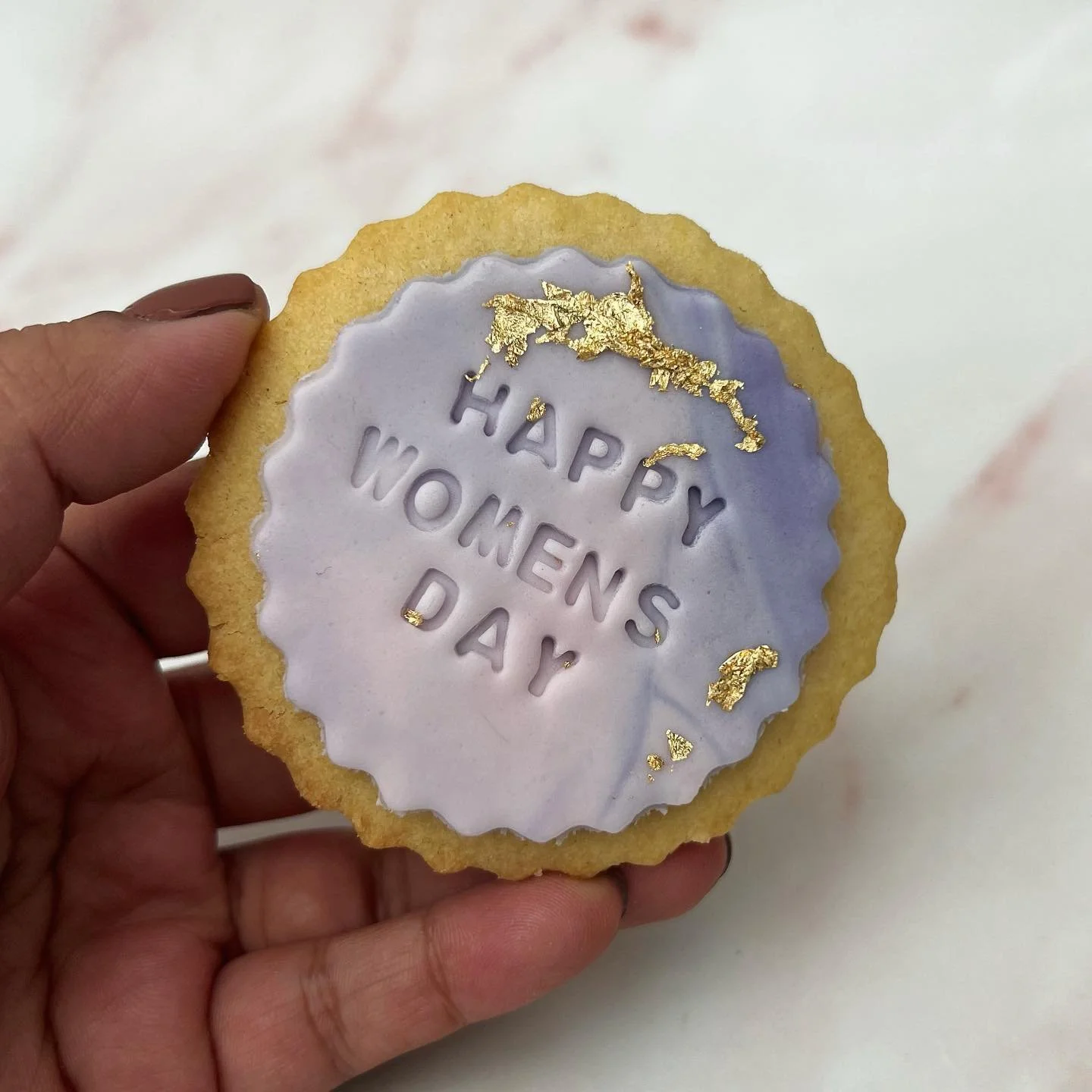 Hand holding a cookie with purple frosting that reads 'Happy Women's Day' and gold leaf decoration.