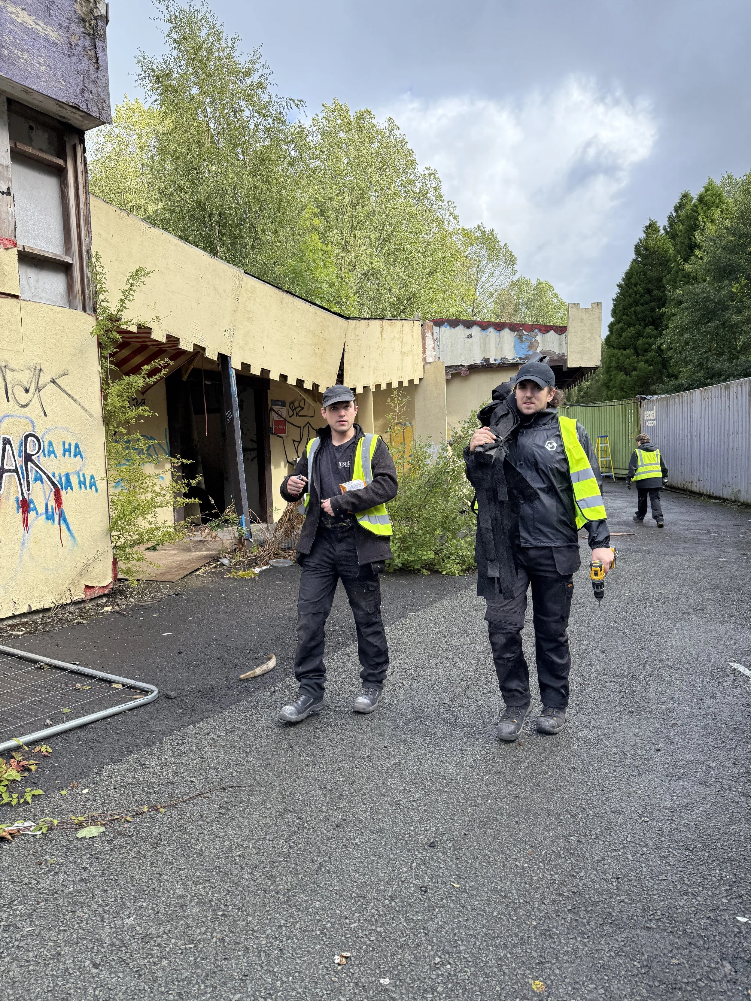 Two young men wearing black clothing and yellow safety vests standing in front of a partially destroyed building with graffiti, surrounded by trees and construction barriers, with another person further back.