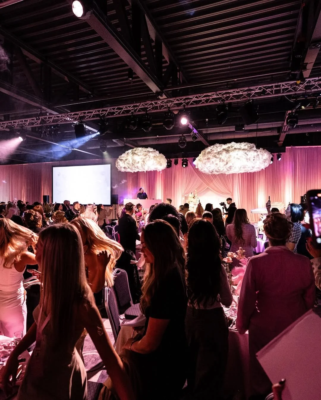 People attending an event with pink drapery, cloud-shaped decorations hanging from the ceiling, and a stage with a speaker and a musician, illuminated by pink and purple lighting.