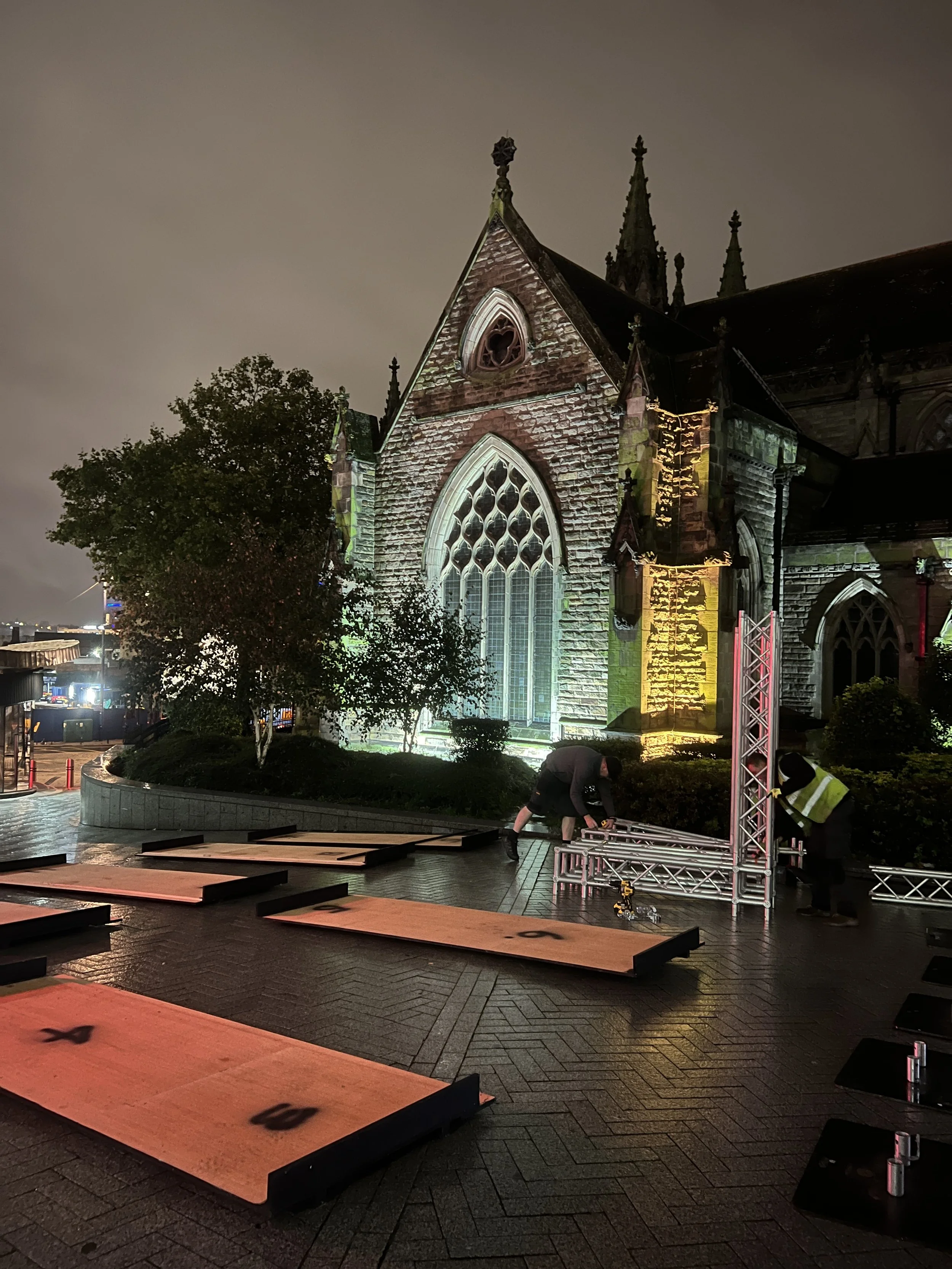 Nighttime photo of a church exterior being illuminated with colorful lights, with workers assembling scaffolding or stage equipment in the foreground, and a tree to the left.