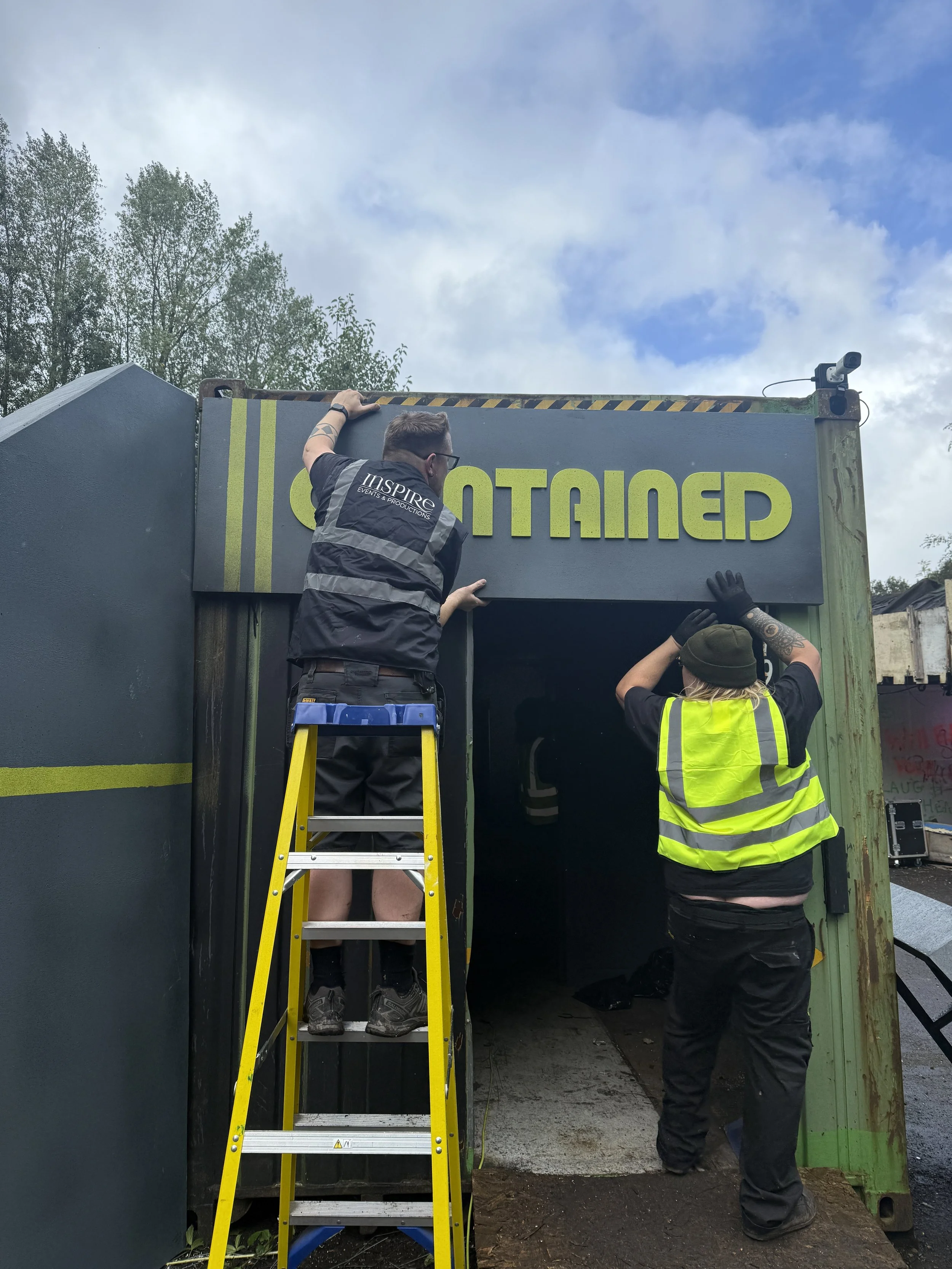 Two workers are installing a signboard. One person is on a yellow ladder, and both are handling the sign, which displays the partially visible word. A security camera is mounted at the top-right corner of the structure, and the background shows trees