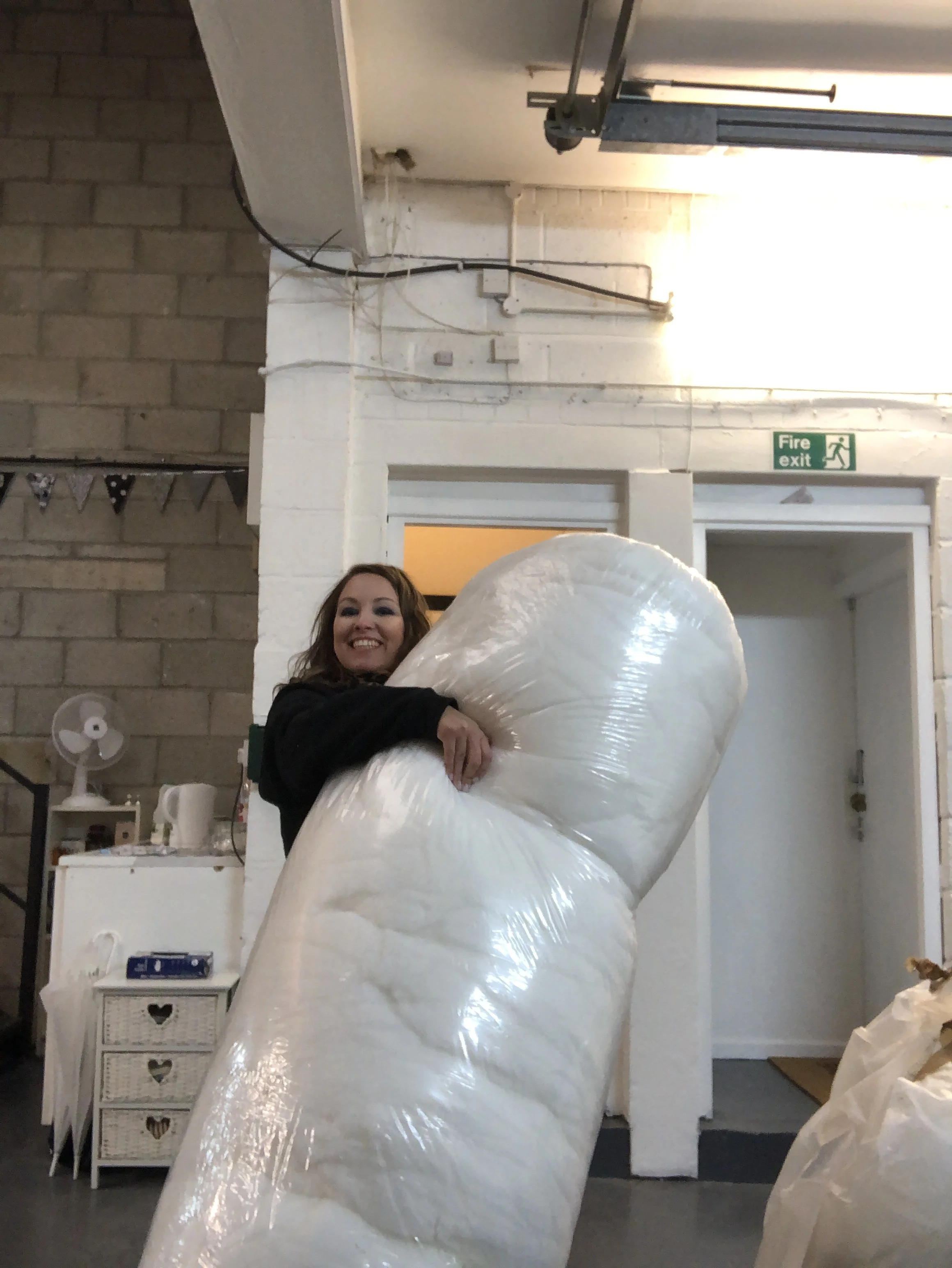 A woman smiling and holding a large roll of plastic wrap in a room with shelves, a white cabinet, a fan, and an emergency exit sign.