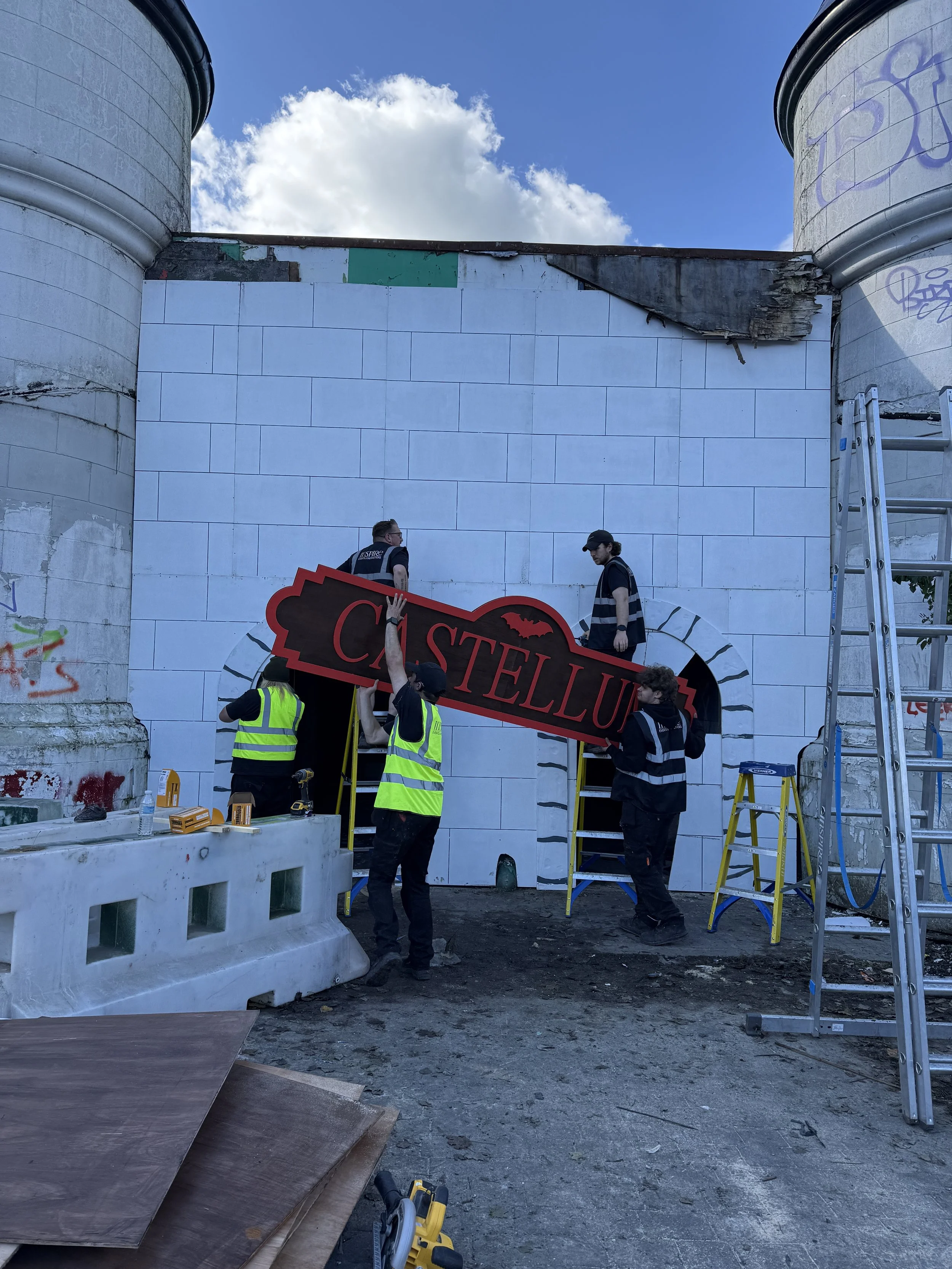 Construction workers installing a sign that says 'Castellum' at the site of a building with graffiti on the walls and a blue sky with clouds overhead.