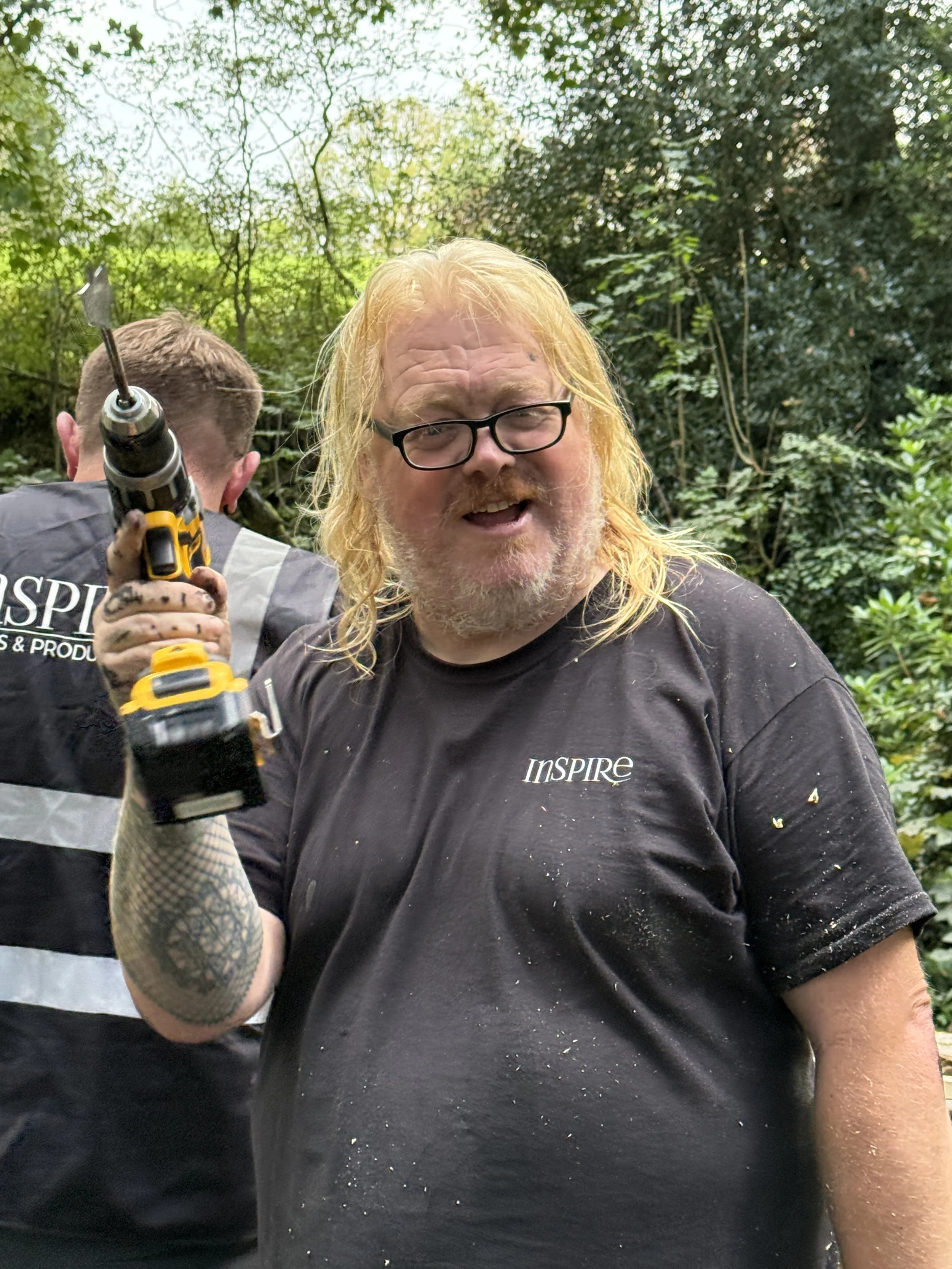 A man with long blonde hair, glasses, and a beard, wearing a black t-shirt with the word 'INSPIRE' on it, holding a power drill, and smiling. There is a person in the background, and foggy green trees in the background.