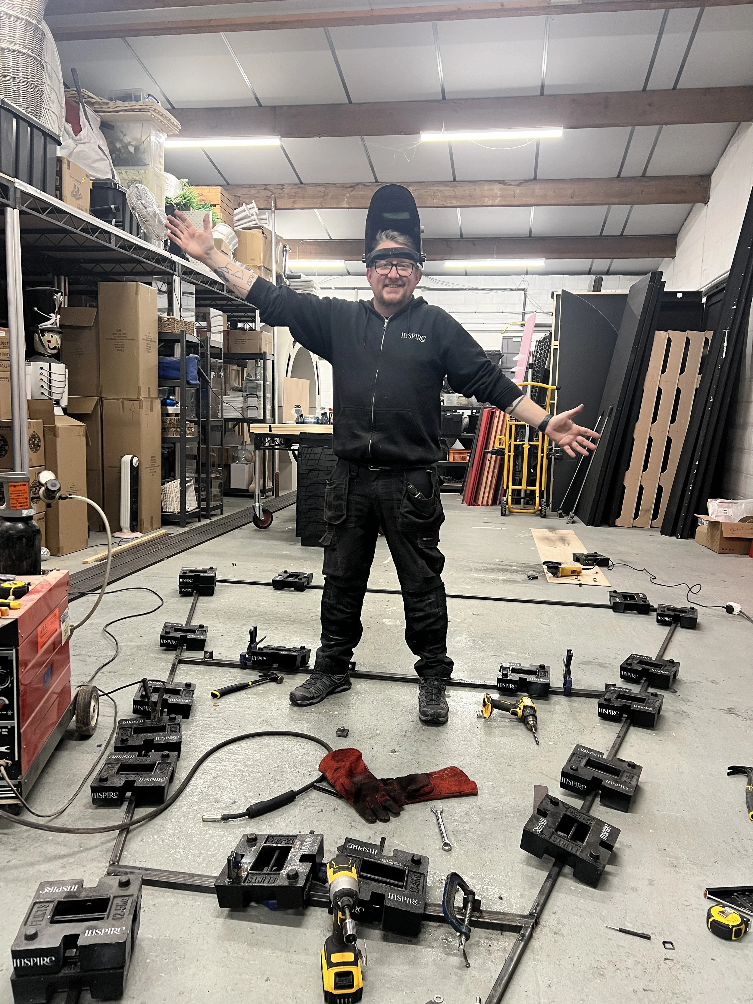 A man wearing a welding helmet on his head and glasses is smiling with arms open in a workshop or warehouse. The floor is scattered with tools, black metal frames, and black boxes labeled Australian Inspire. Shelves behind him contain boxes, pipes, a