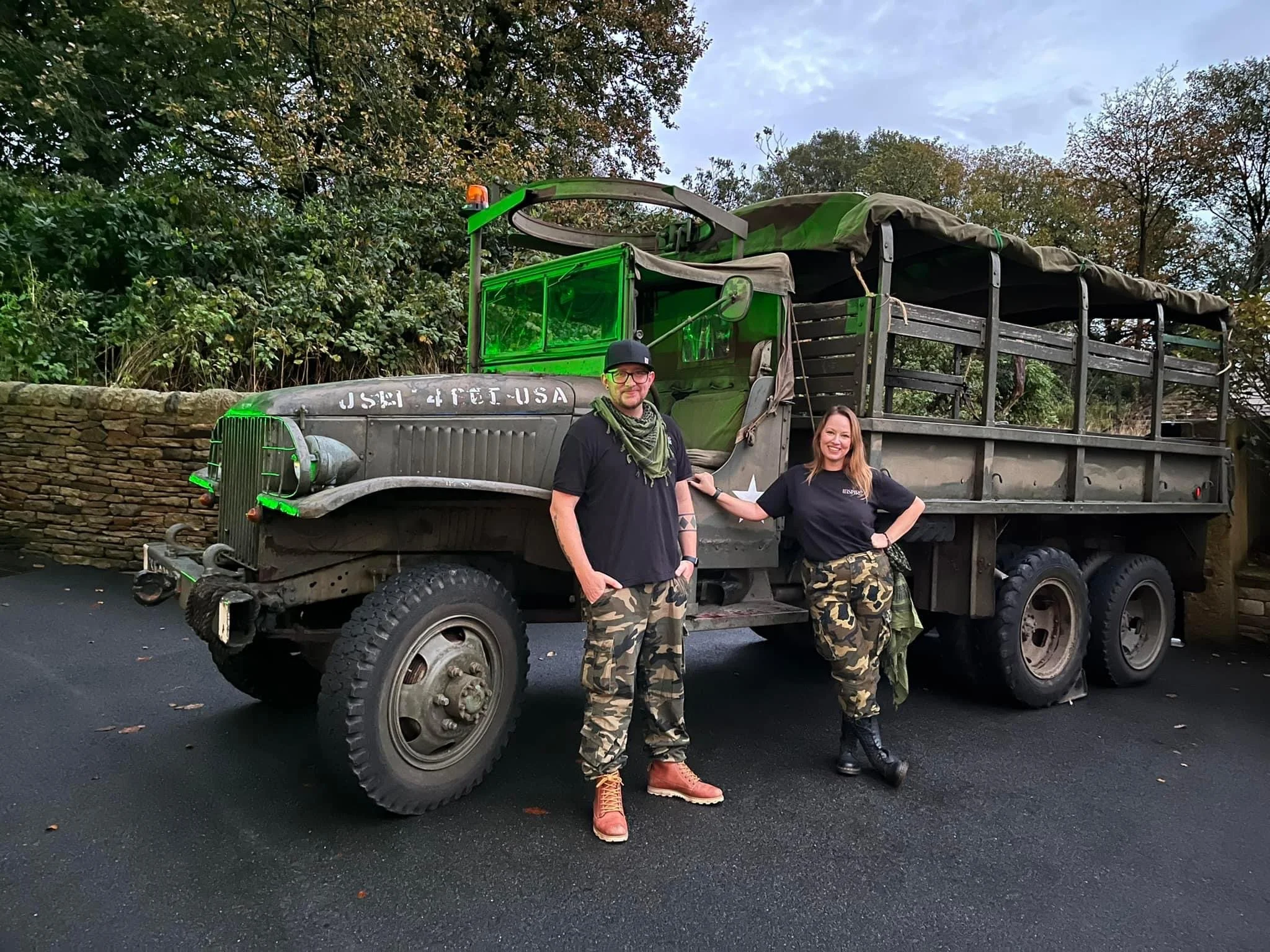 Two people standing and smiling next to a military truck with a green cargo cover on a paved street with trees in the background.