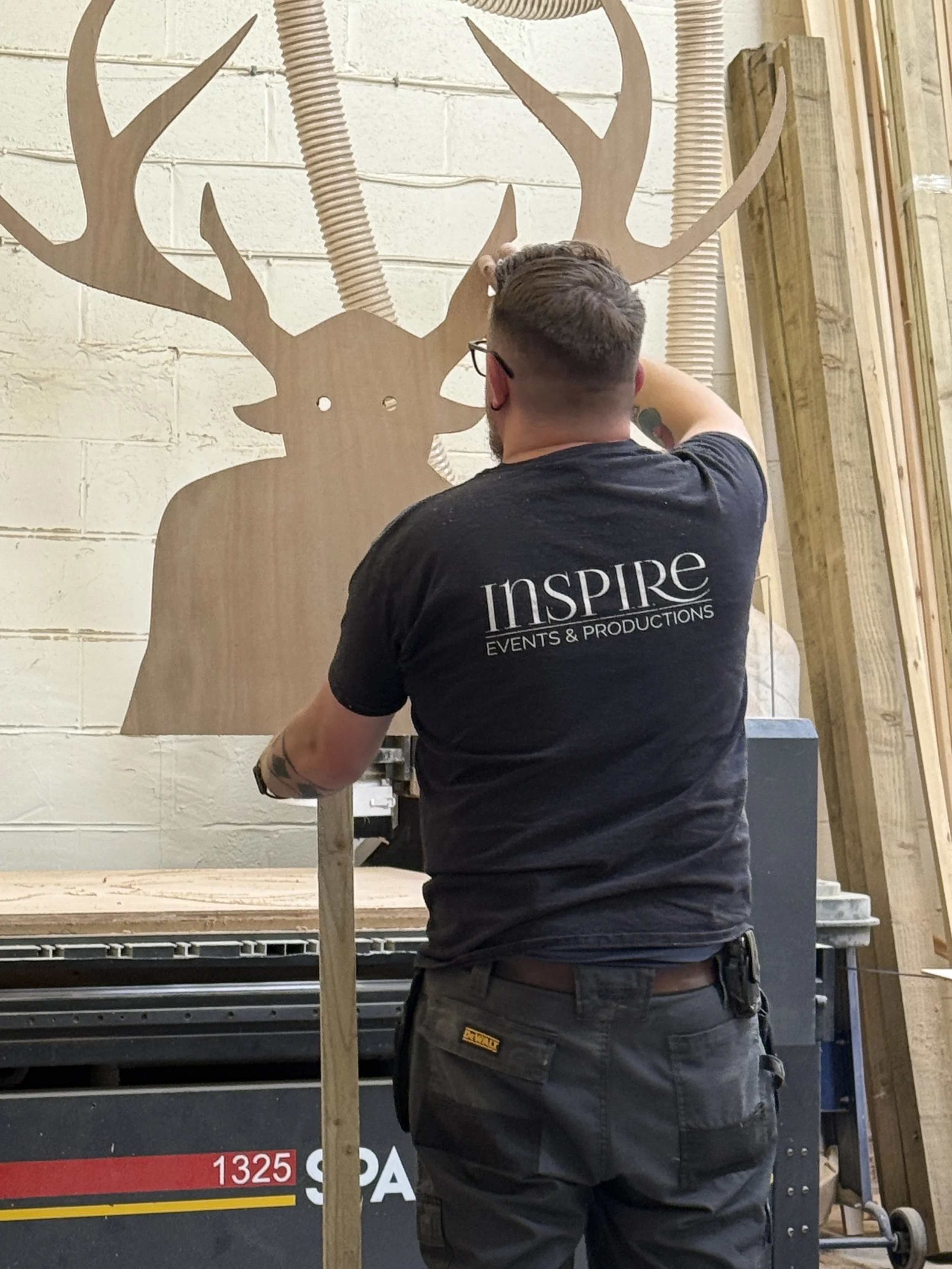 A man working in a woodworking or carpentry shop. He is wearing a black t-shirt with the word 'INSPIRE' on the back and black cargo shorts. He is standing in front of a wooden deer head with large antlers mounted on the wall, and appears to be using a tool on it. The shop has wood planks and equipment in the background.