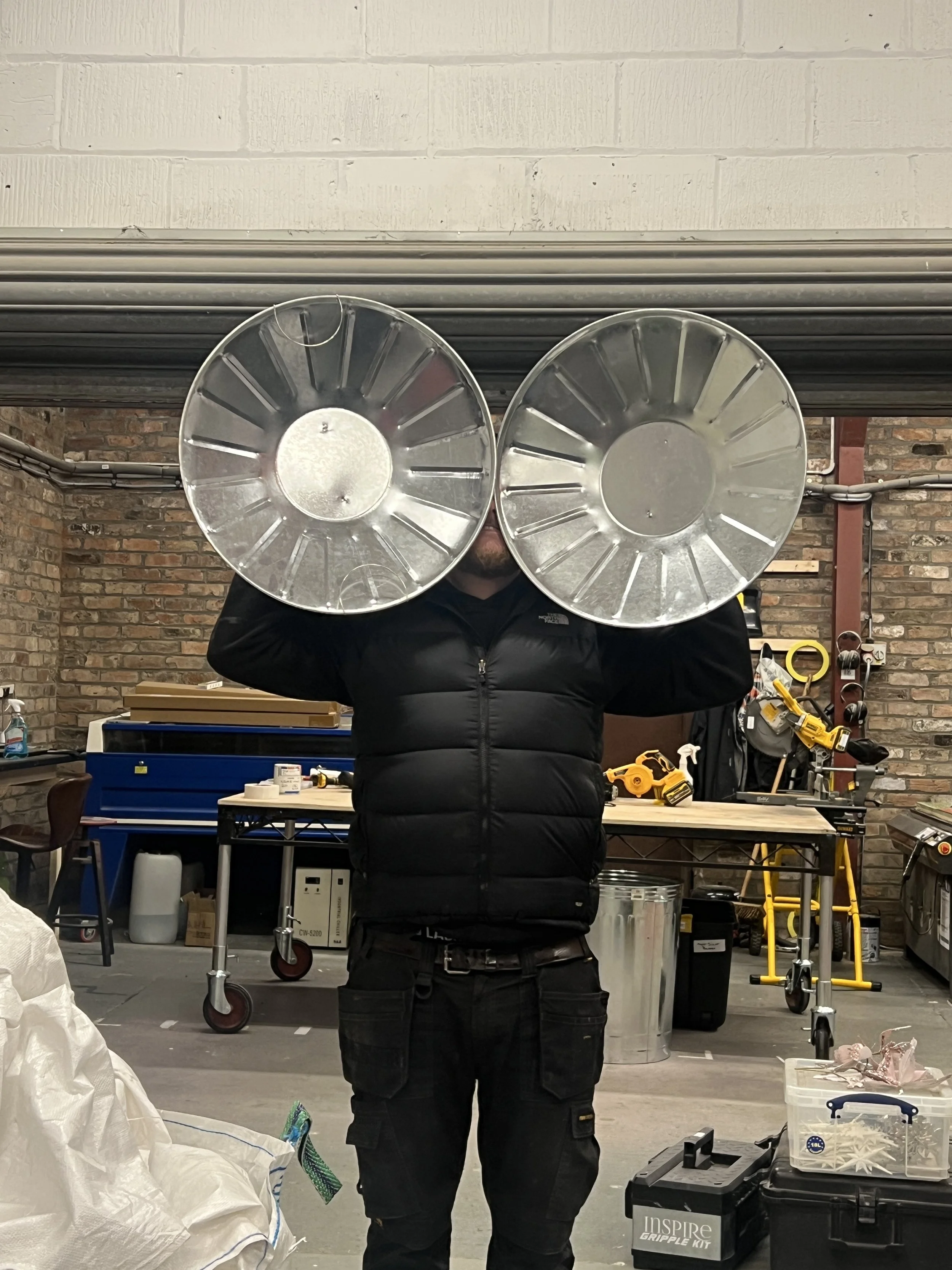Person holding two large metal reflective dish-shaped objects in a workshop with tools, storage boxes, and brick walls in the background.