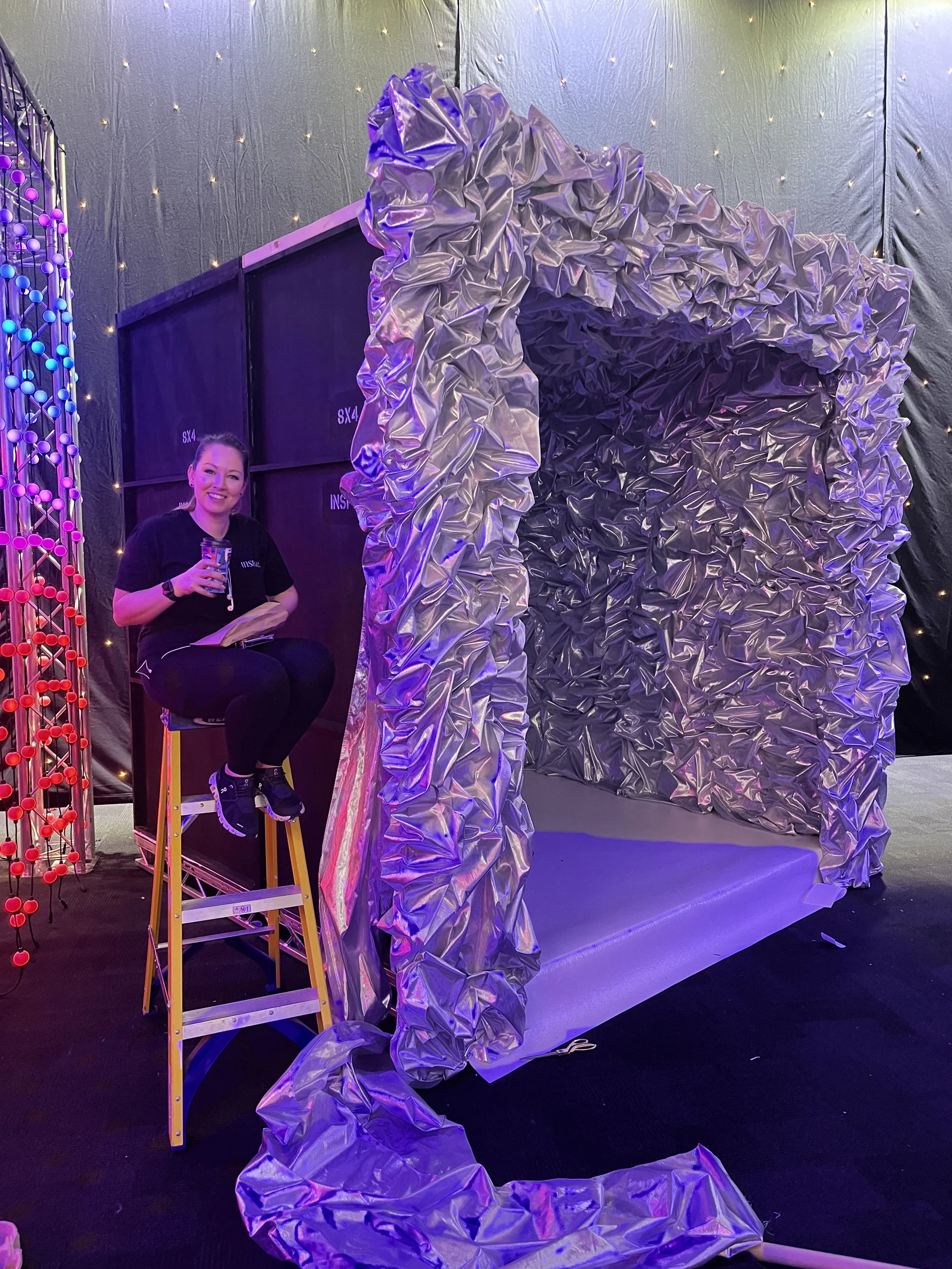 A woman sitting on a ladder, smiling and holding a drink, next to a large, crumpled metallic structure, with colorful LED lights and a black backdrop.