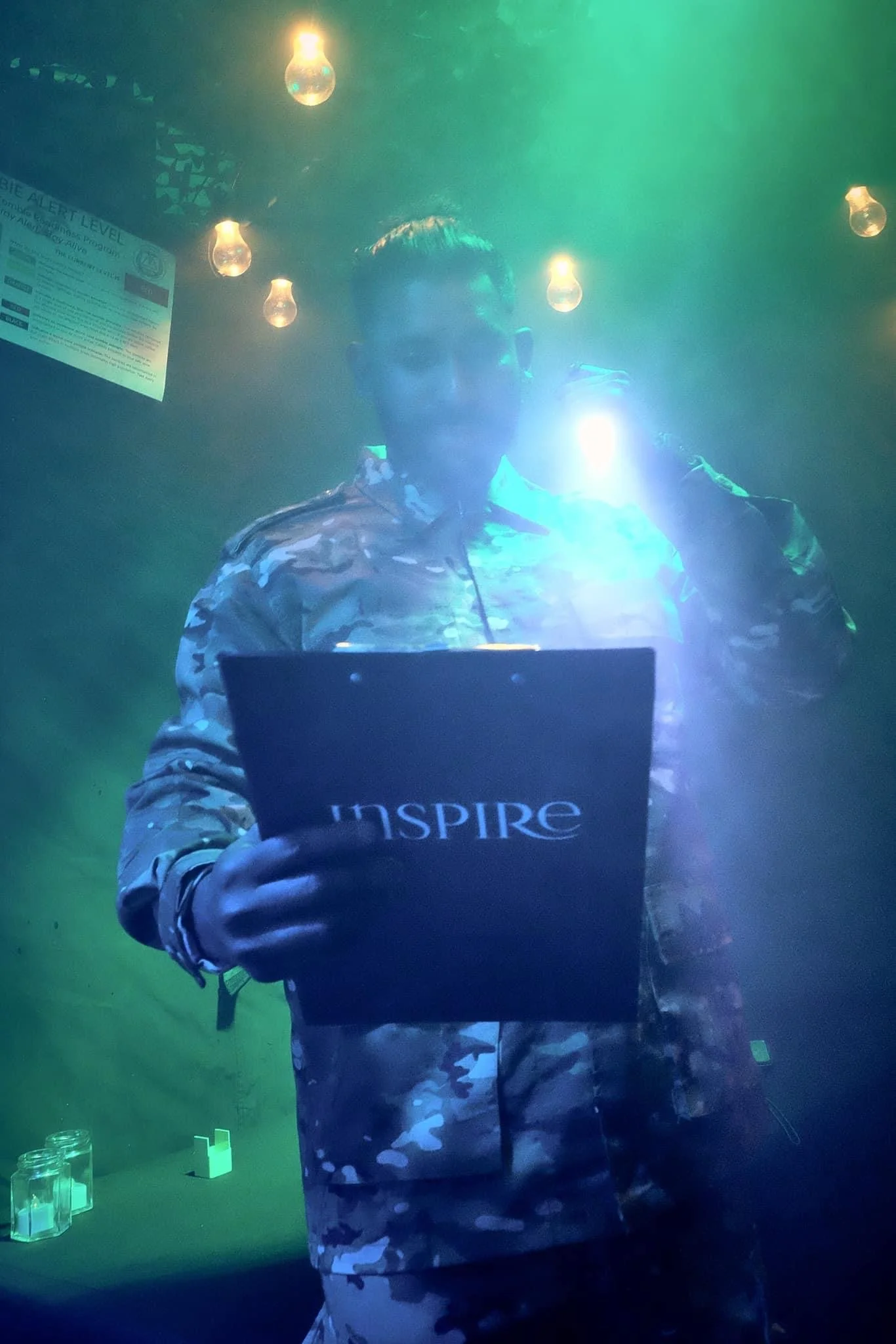 A person taking a mirror selfie at a dimly lit bar or club, wearing a camouflage jacket and holding a book or sign that says 'Inspire'. The background features hanging light bulbs and a screen displaying emergency alert information.