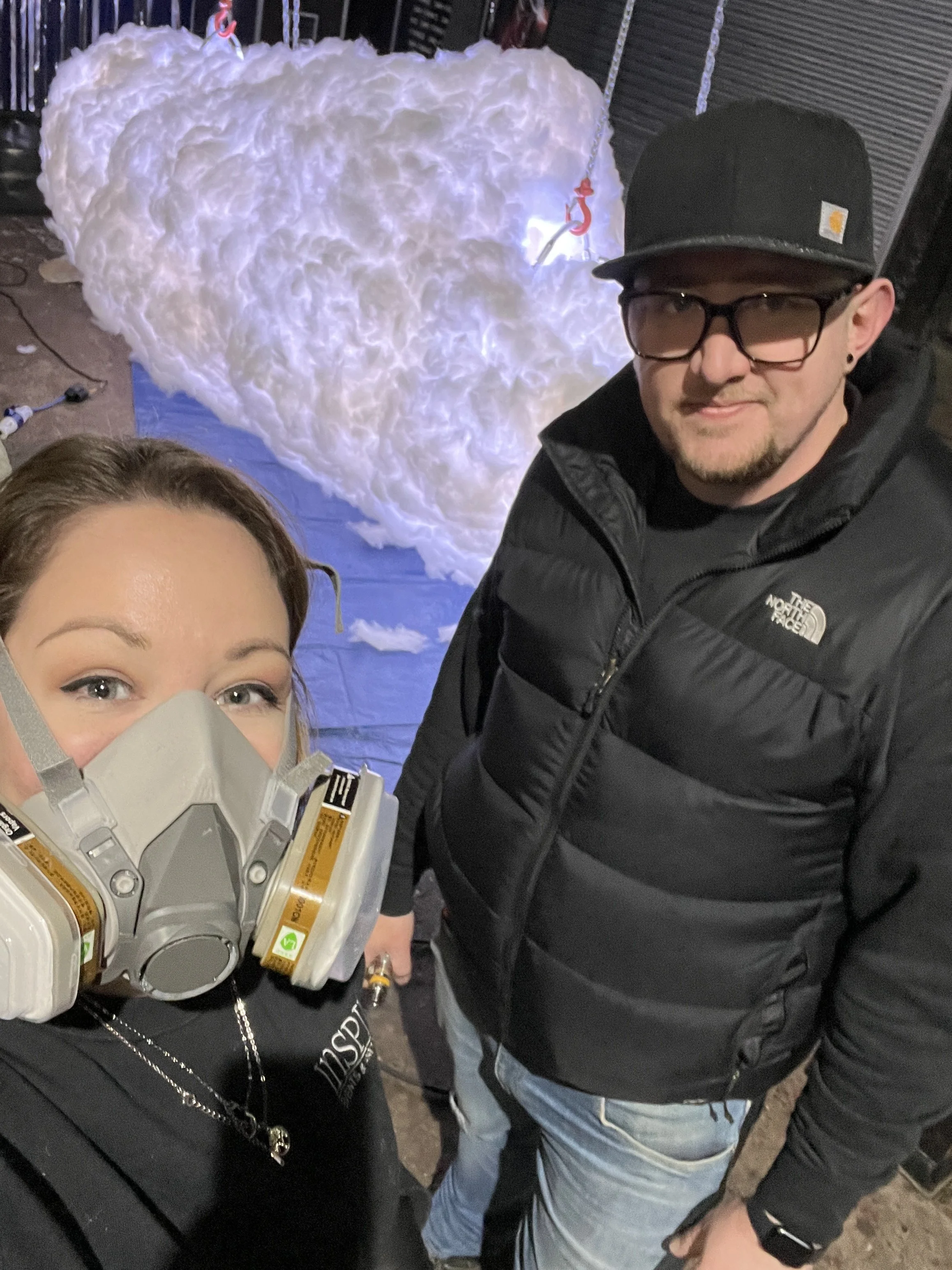 Close-up selfie of a woman with light skin, blonde hair, and blue eyes wearing a face mask. Partially visible in the background are a man with light skin, wearing glasses, a black North Face jacket, and a black cap. Behind them, there is a large hear