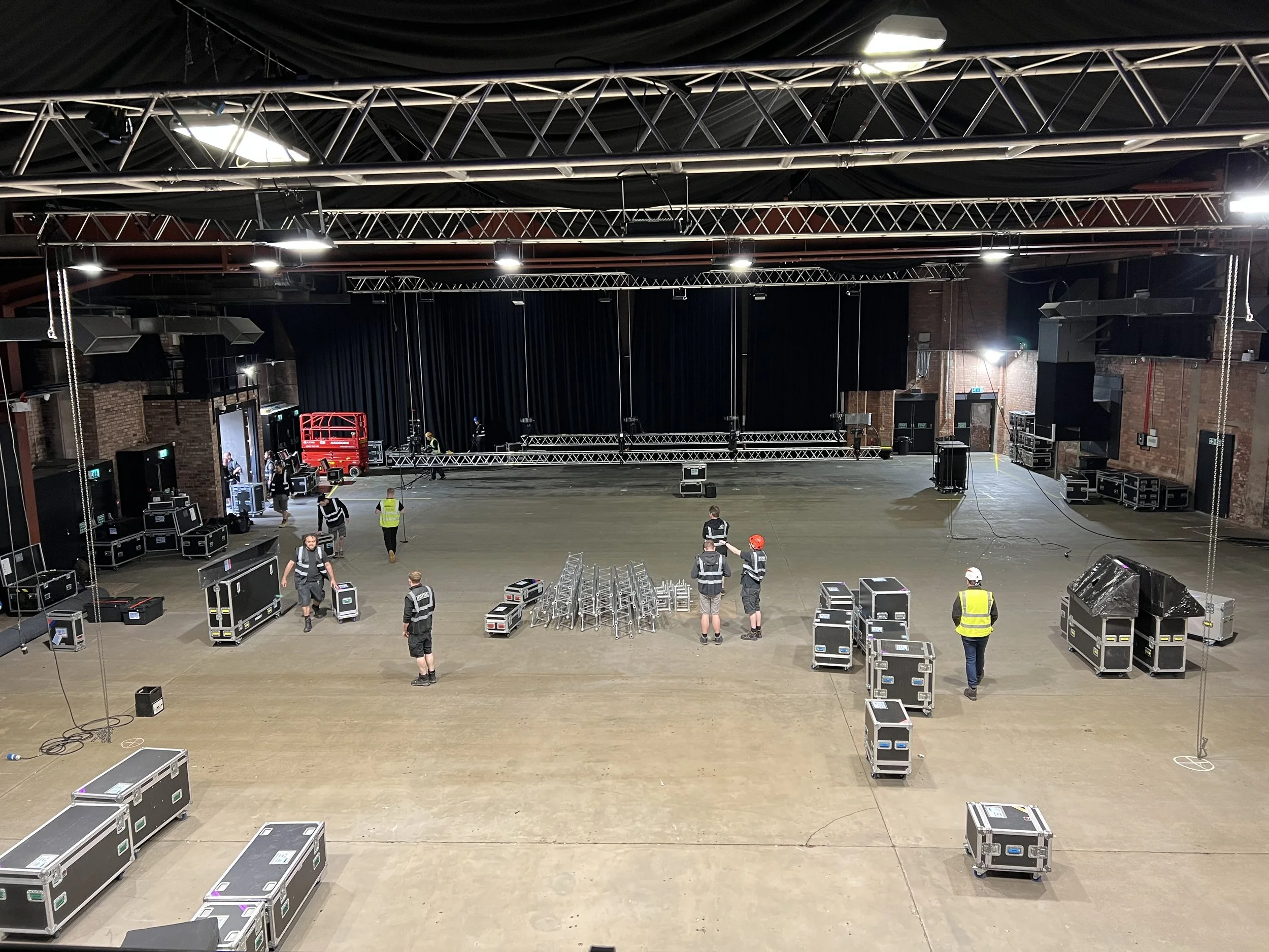 Large indoor venue with stage equipment and lighting being set up, crew members in safety vests working, and equipment cases scattered across the floor.