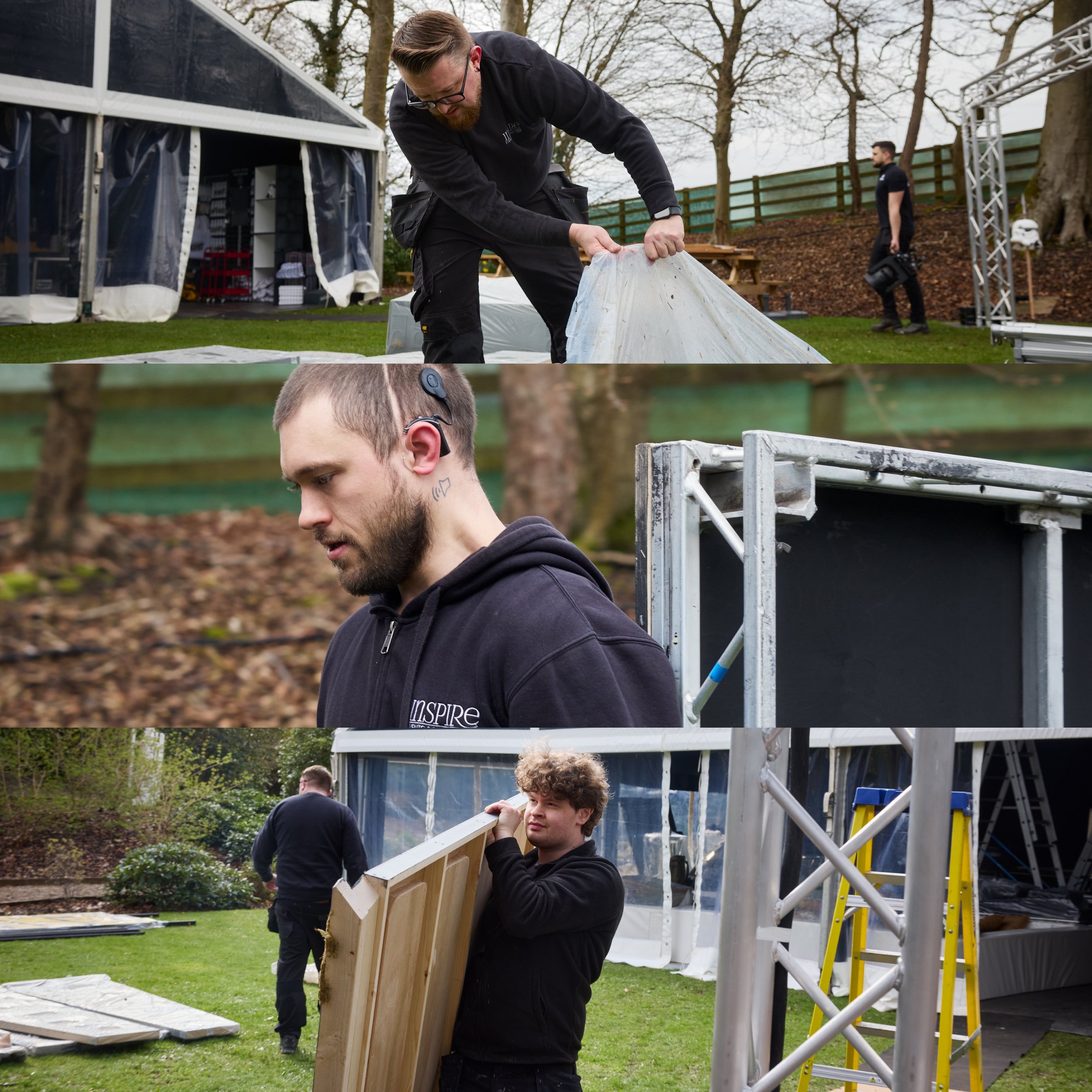 A man with glasses and a beard in black clothing is assembling a wooden structure outdoors during daytime, with a second man in black nearby and trees in the background.