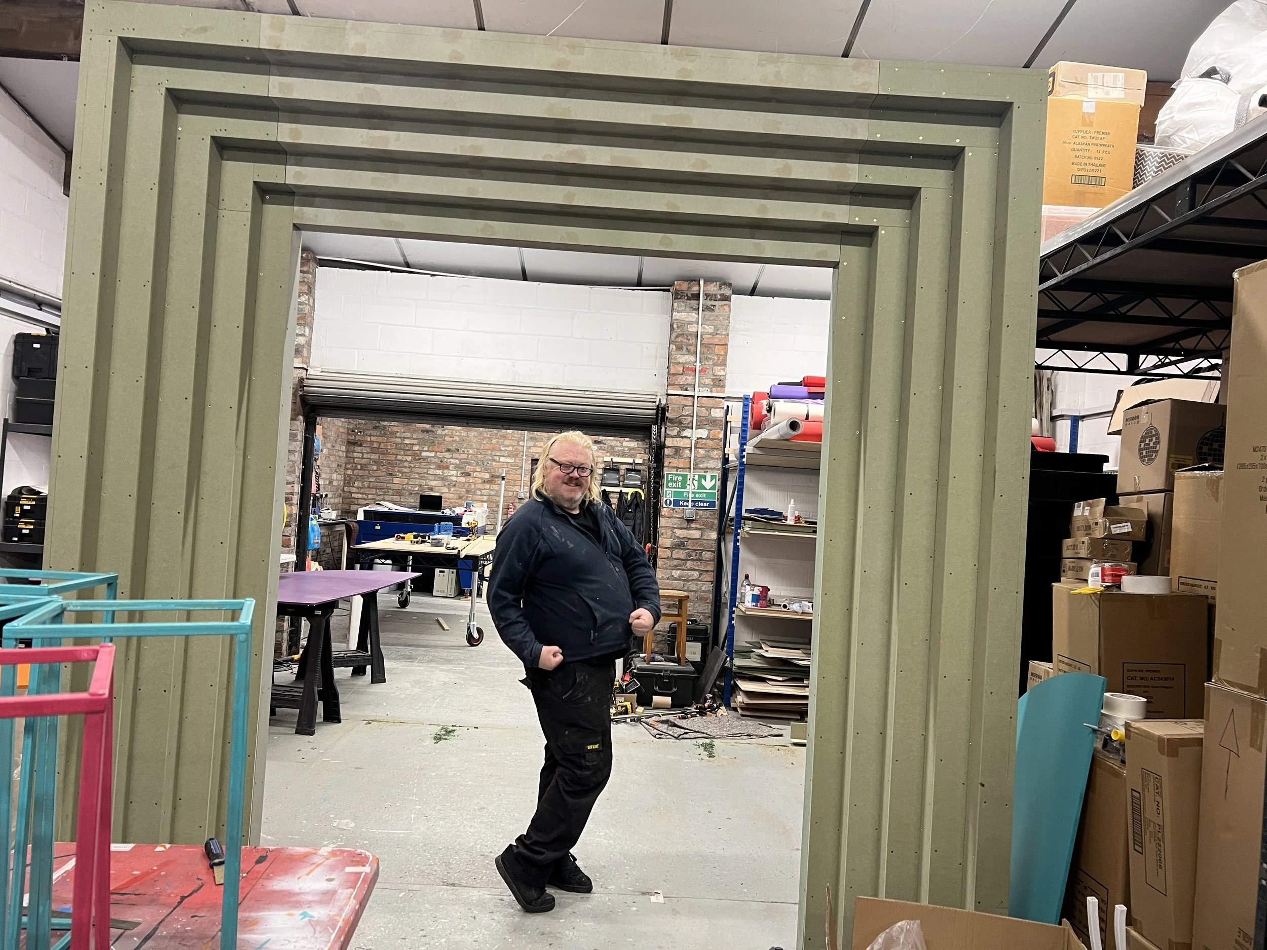 A man with glasses and long blonde hair standing in a workshop, smiling, within a green metal frame structure, surrounded by various tools, boxes, and shelves.