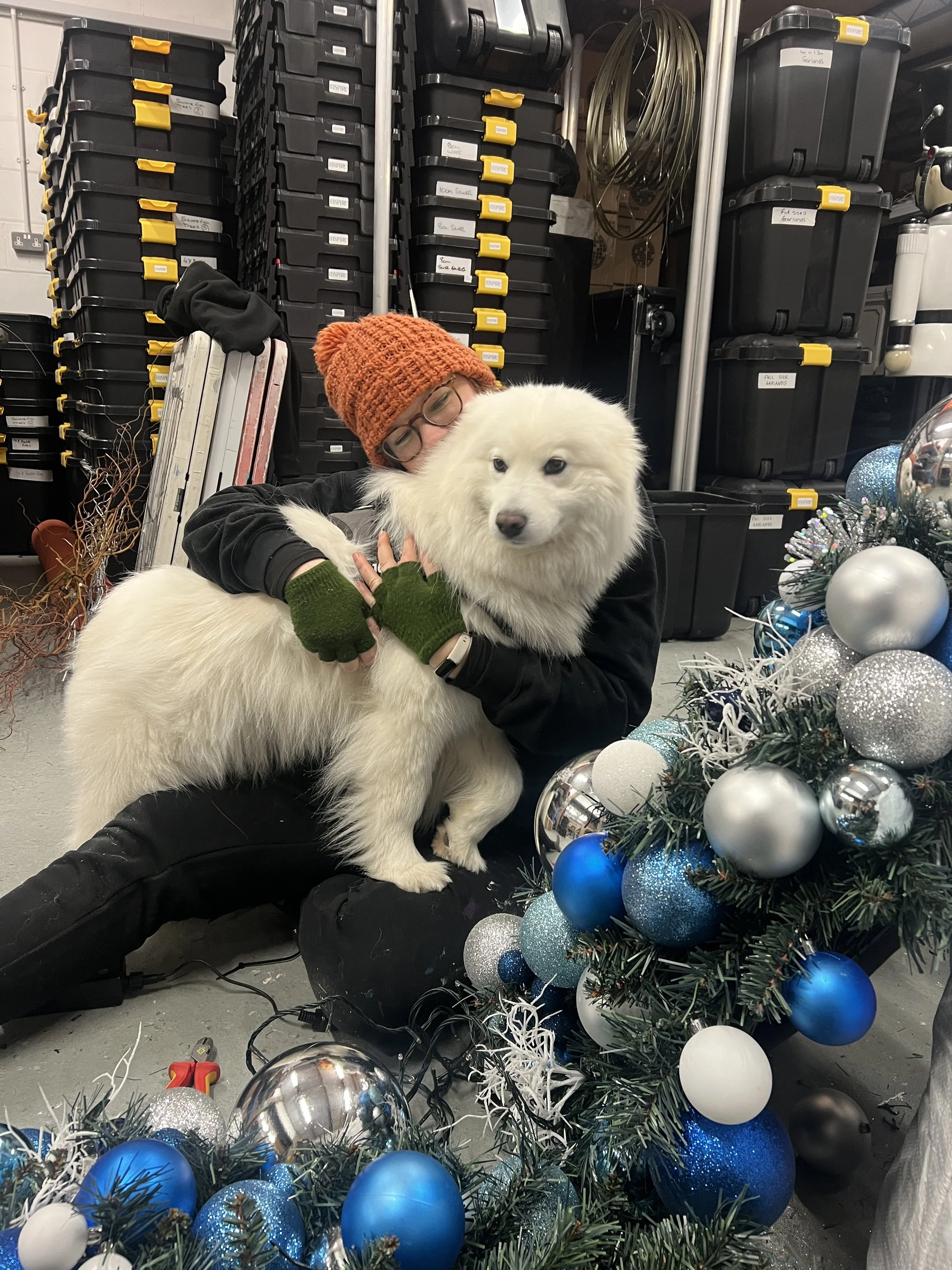 A person with glasses, wearing a knitted orange hat, hugging a fluffy white dog in an industrial setting with stacked storage bins and various decorations around.