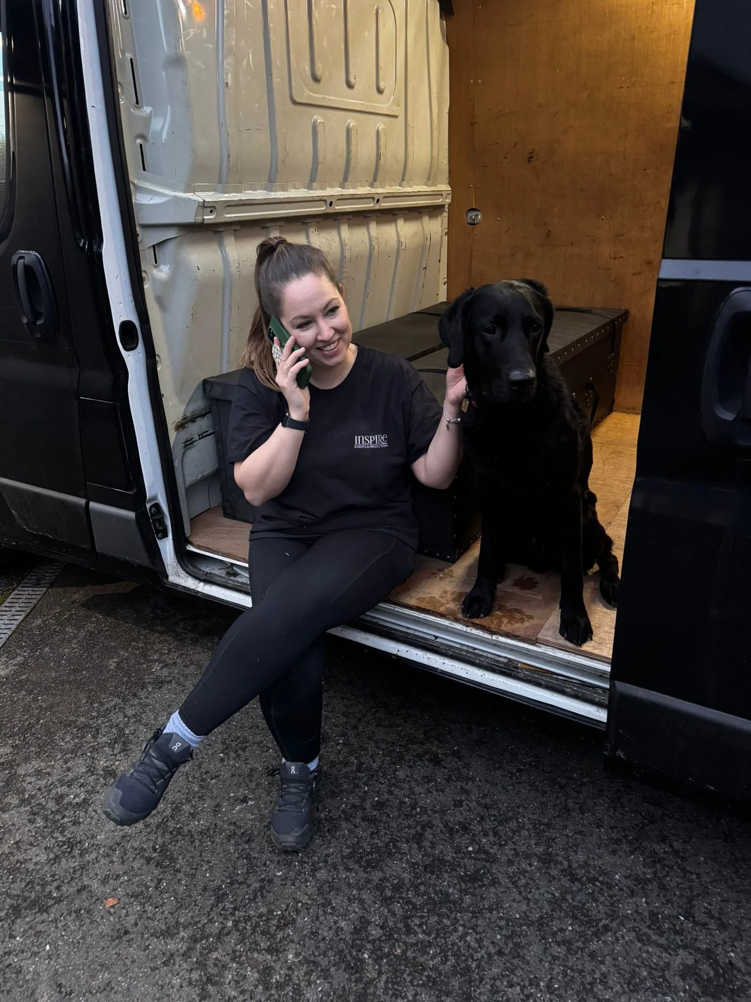 A woman sitting on the edge of a van with a large black dog. The woman is talking on a phone, smiling, and wearing black athletic clothing and sneakers. The van's interior shows the walls and some equipment or storage inside.