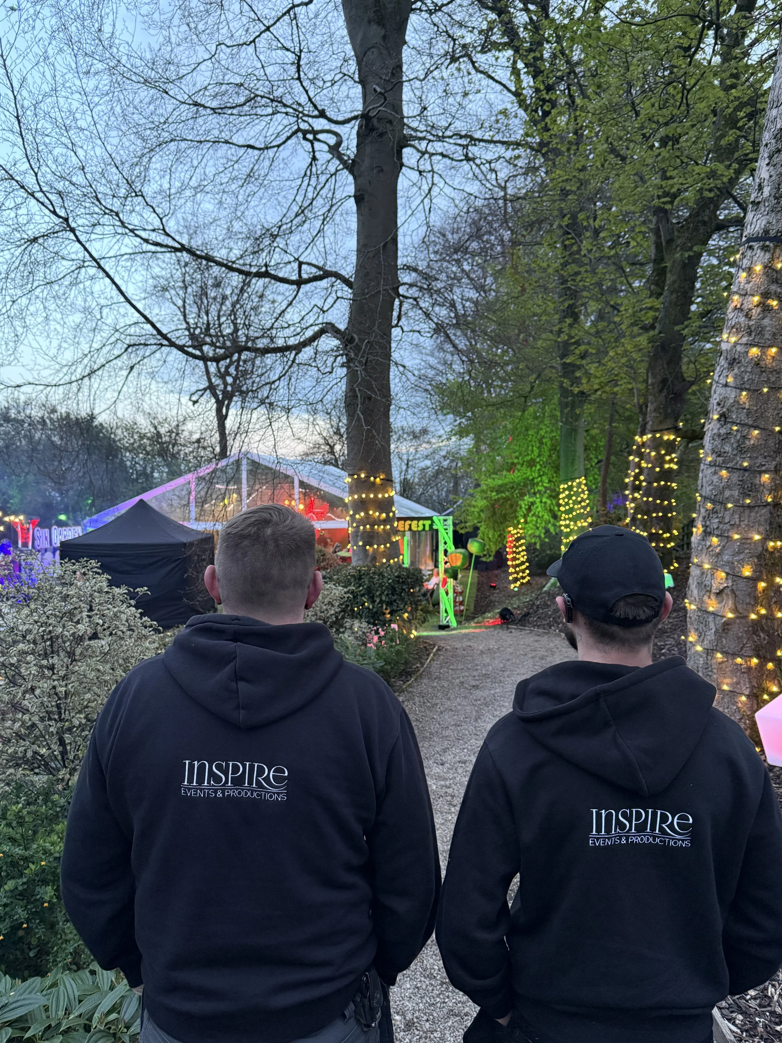 Two men wearing black hoodies with 'Inspire Events & Productions' logo, standing outdoors with trees decorated with string lights, and a festive event area visible in the background.