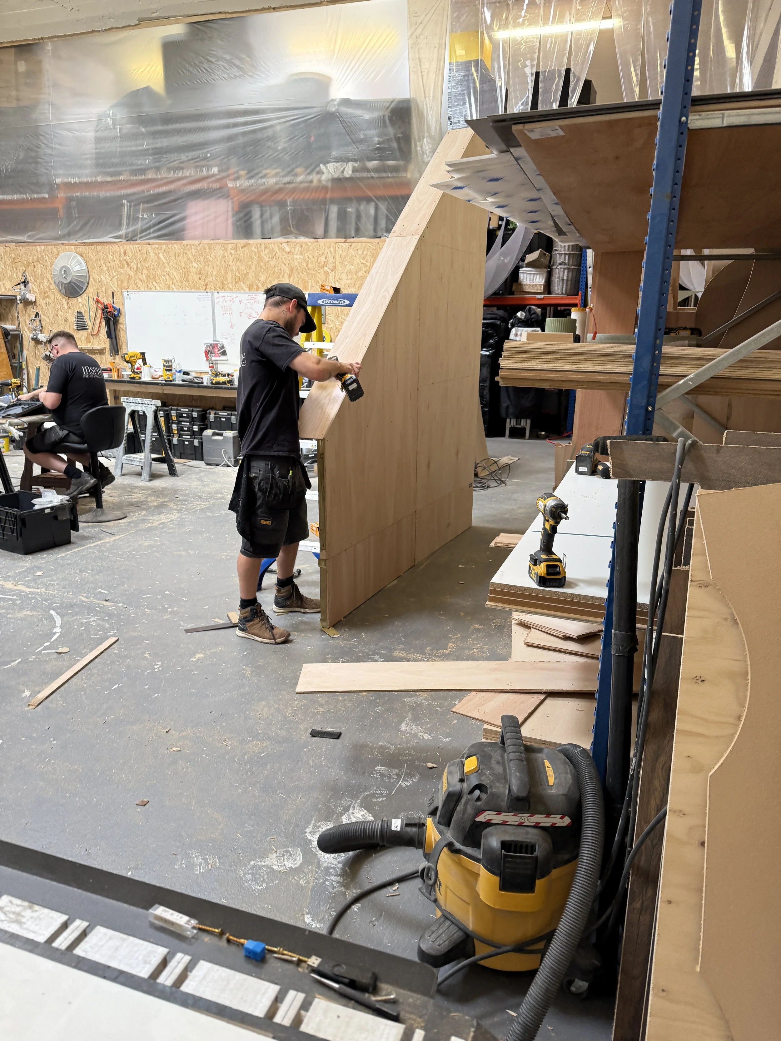 Two men working on constructing a wooden structure in a workshop filled with tools and supplies.