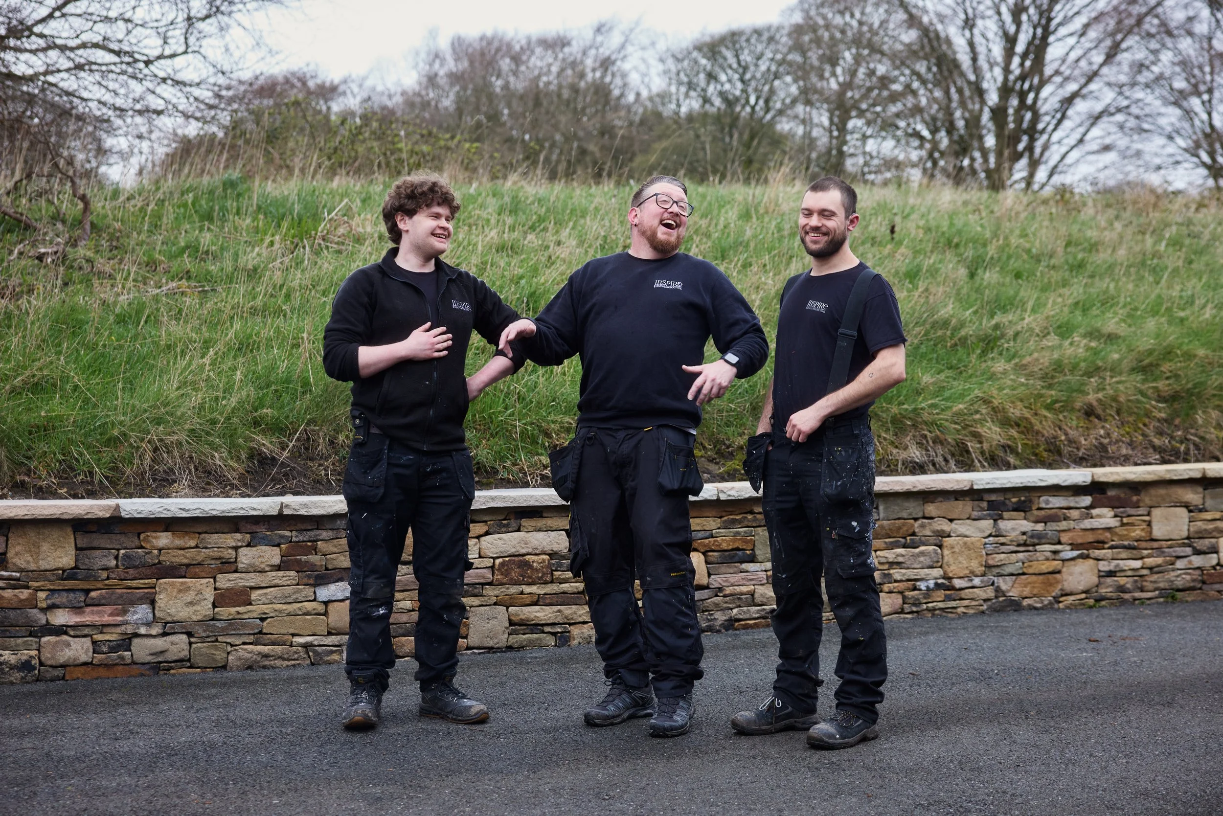 Three men in work clothes, standing outdoors on a paved surface, smiling and laughing together, with a grassy hill and leafless trees in the background.