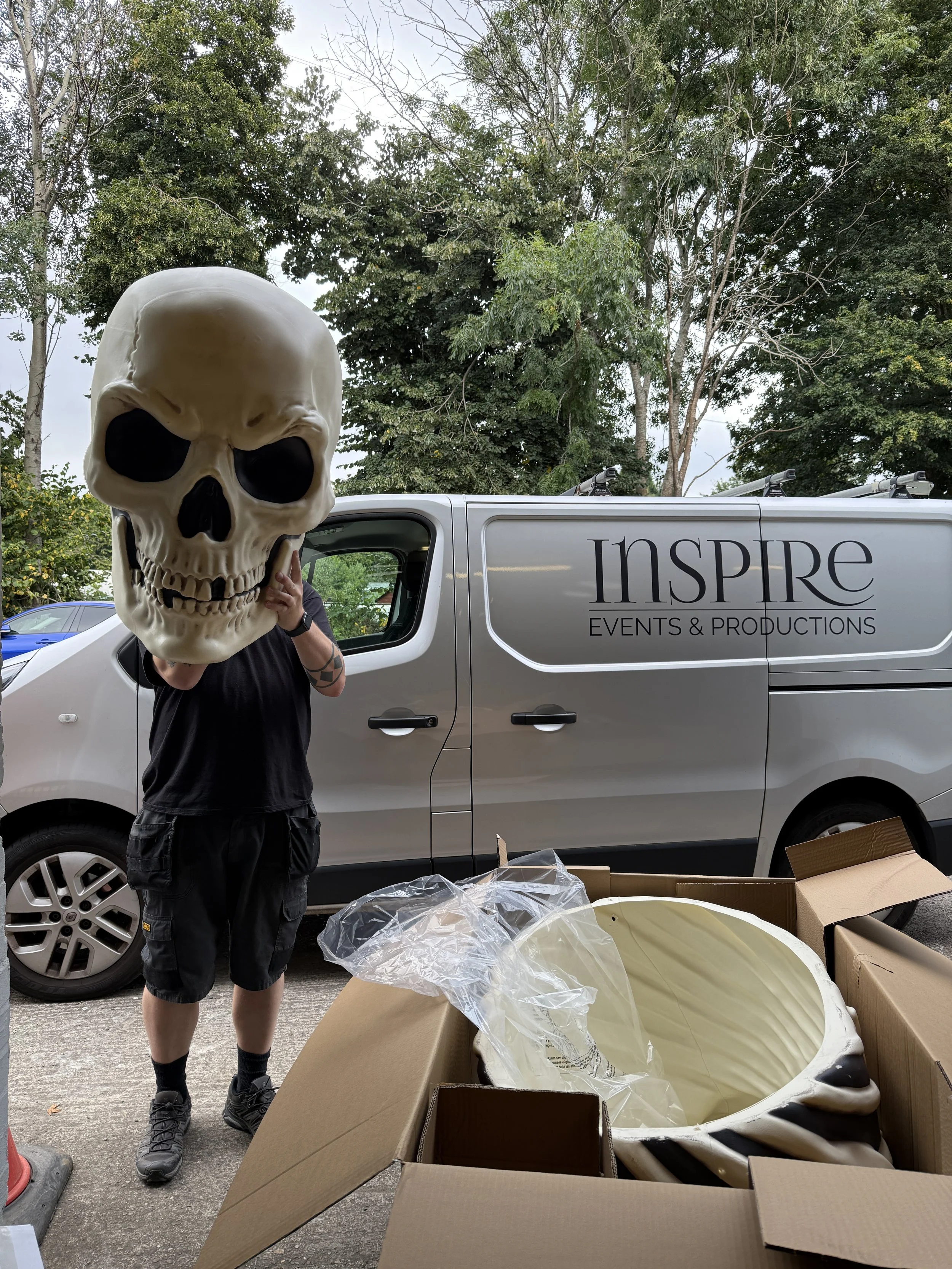 Person holding a large skull mask in front of a vehicle with the words 'INSPIRE EVENTS & PRODUCTIONS' on it, surrounded by cardboard boxes and plastic wrap outdoors with trees in the background.