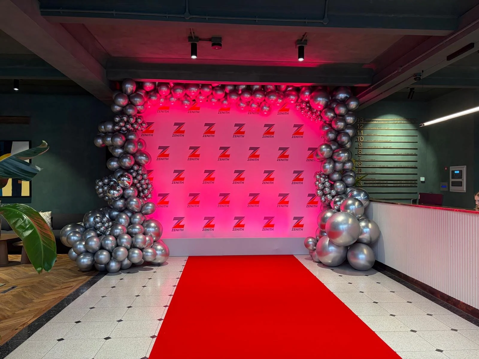 Event backdrop with pink lighting, decorated with silver balloons, and a red carpet leading to it, in an indoor venue.
