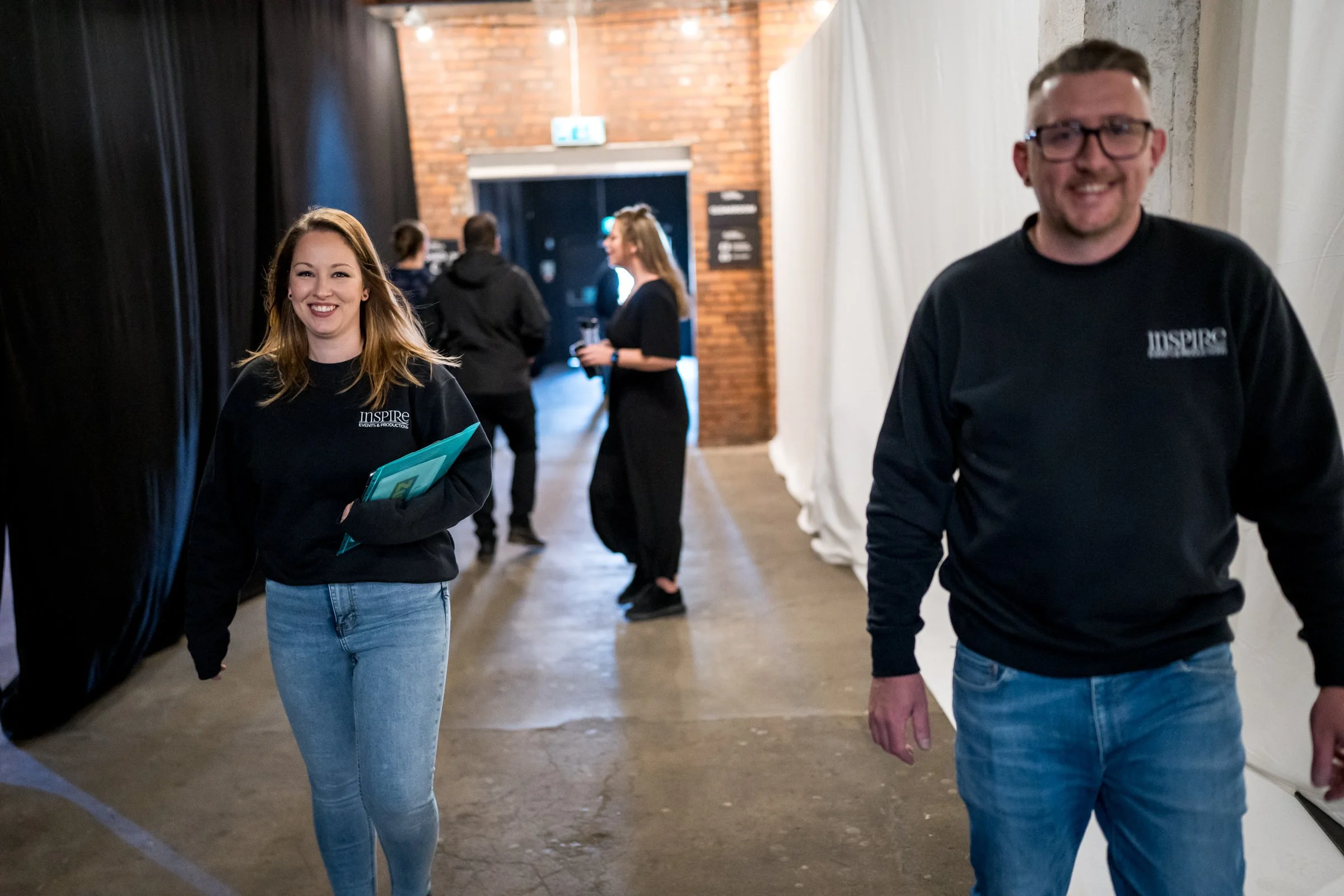 People walking in a hallway with brick and white walls, some engaged in conversation, wearing black shirts with 'INSPIRE' logo.