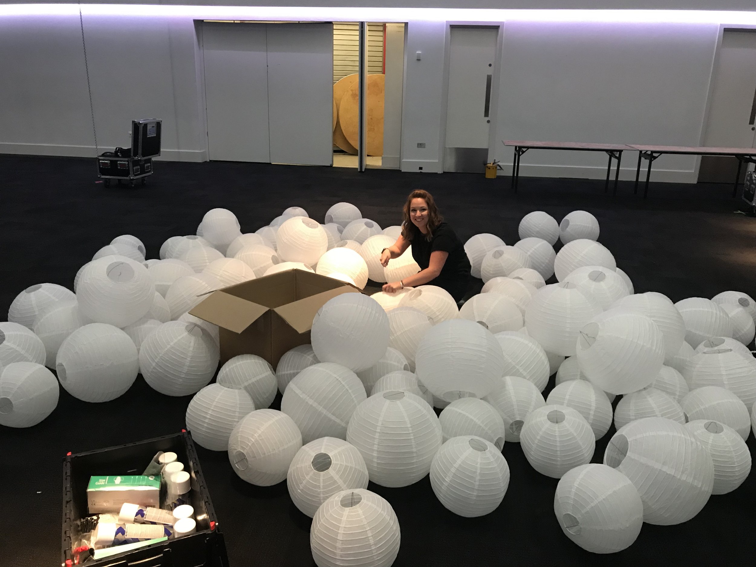 A woman sitting among white paper lanterns in a room with a black floor, with tables and a door in the background.