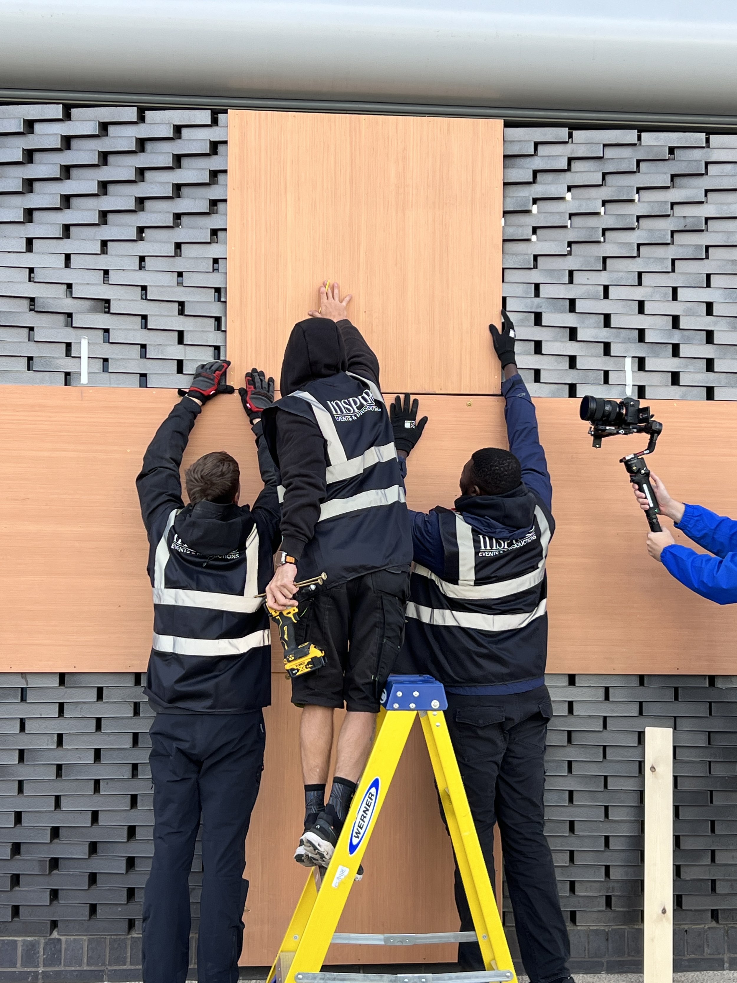 People working together to hang a large wooden panel on a dark brick wall, one person on a yellow ladder, others supporting the panel from below, with a person holding a camera stabilizer on the right.