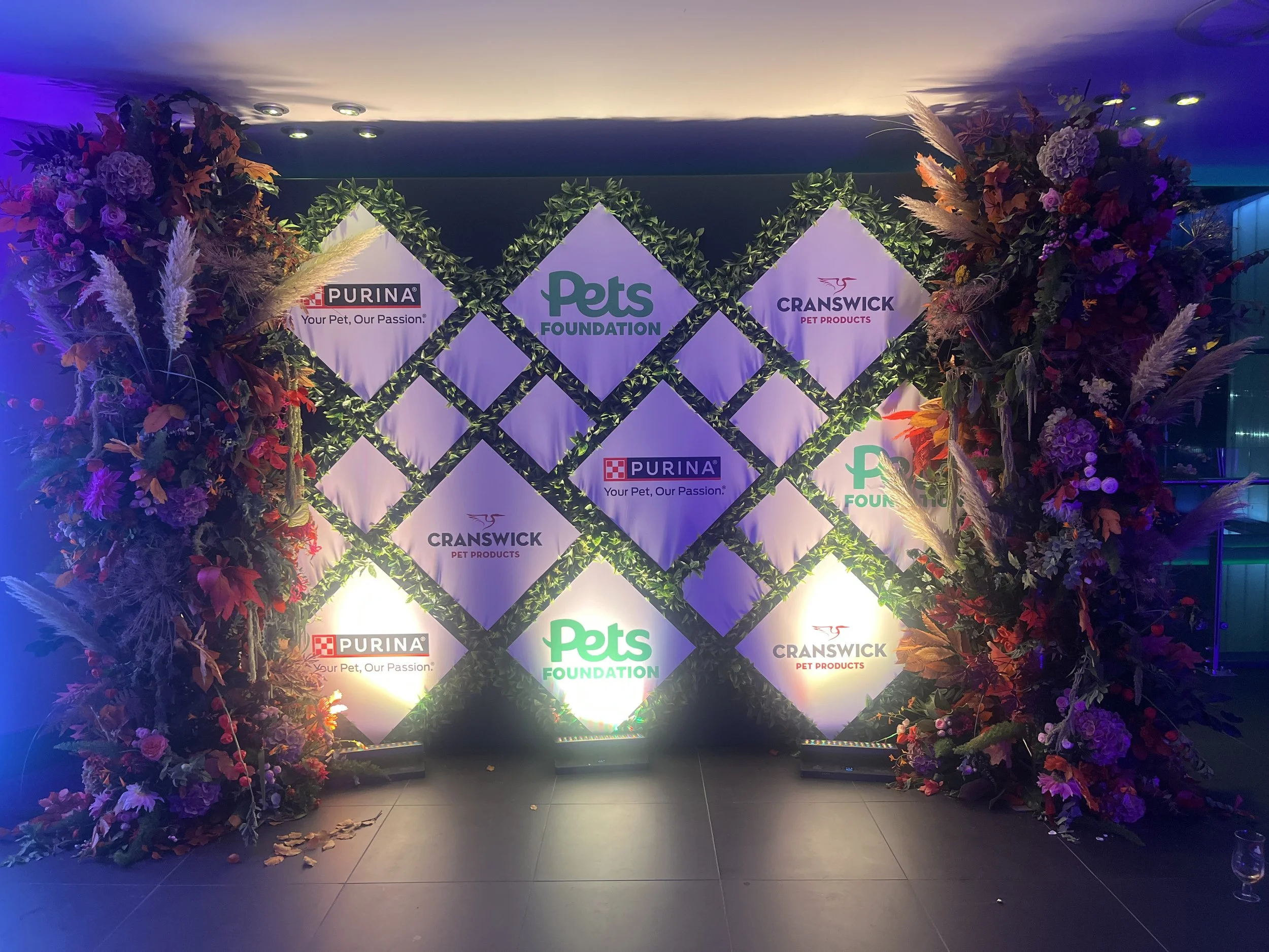 Decorative backdrop with diamond-shaped logos of Purina, Pets Foundation, and Cranswick Pet Products, surrounded by colorful floral arrangements and pampas grass.