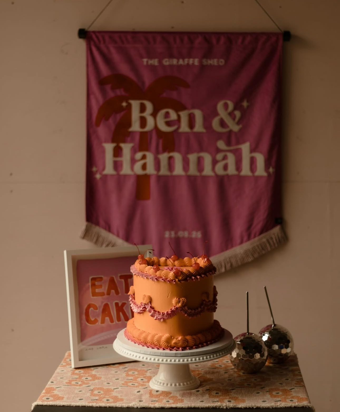 A lambeth vintage style heart shape orange and pink cake with decorations on a white cake stand. Behind the cake is a framed sign that reads 'EAT CAKE,' and two disco ball decorations with straws. 