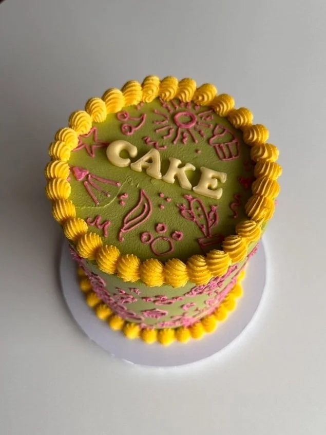 A round cake with green and pink frosting, decorated with the word "CAKE" in white letters on top, and personalised squiggle drawings to represent the birthday person, a yellow piped border around the edge.