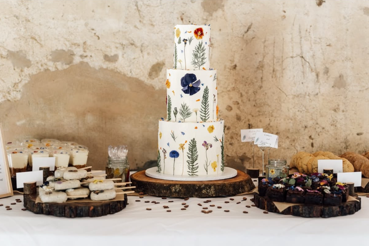 Three-tiered wedding cake with pressed edible flower design, placed on a wooden slab, surrounded by cookies, brownies, and mini vegan cheesecakes on a white tablecloth.
