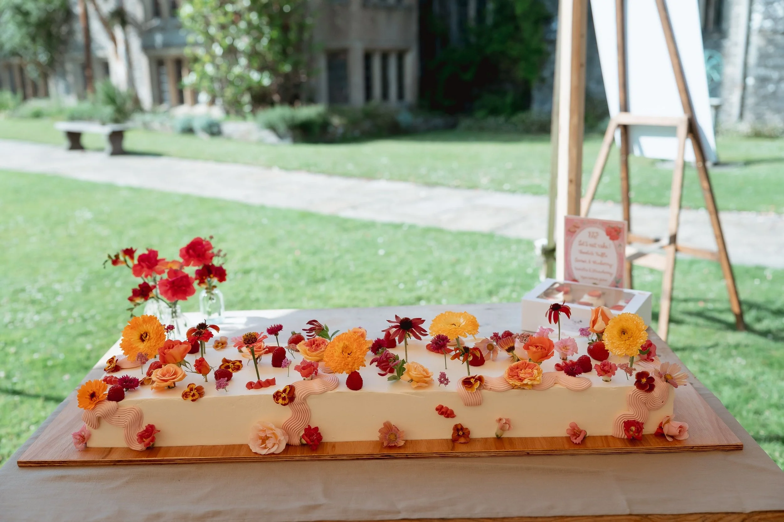 A rectangular white wedding cake decorated with pink and orange flowers, including roses and other blooms, and pink icing accents, placed on a wooden board on a table outside at a garden wedding.
