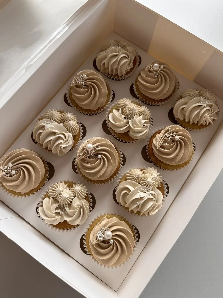 A white box filled with twelve decorated cupcakes, each with swirled beige and white frosting, some topped with pearl-like sprinkles and small edible beads.