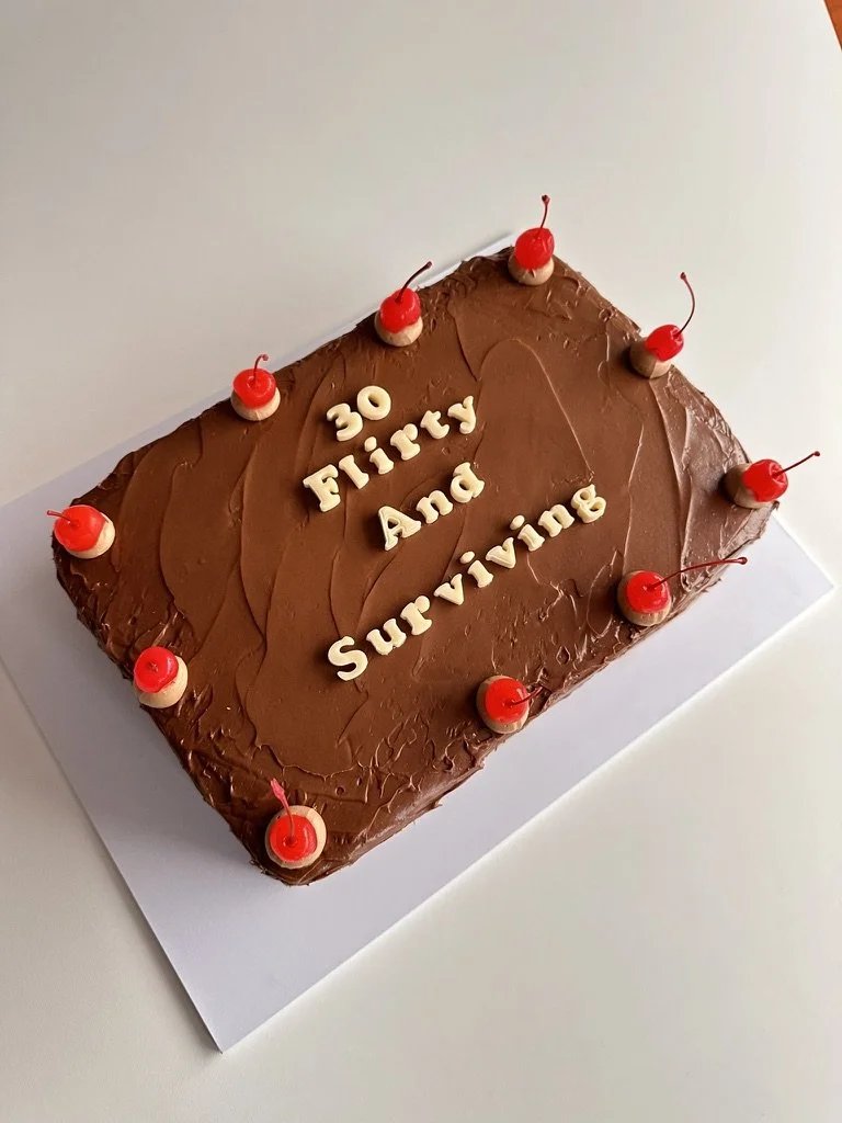 Chocolate fudge birthday cake with fondant letters that says '30 Flirty And Surviving' and decorated with red maraschino cherries around the edges.