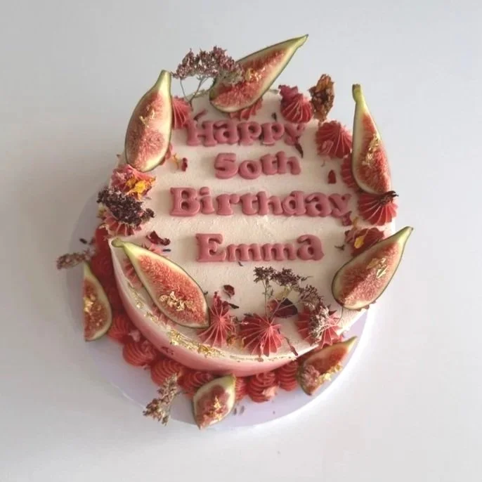 A birthday cake decorated with slices of figs, pink frosting, and small pink and brown decorative flowers. The cake has a message that reads "Happy 50th Birthday Emma".