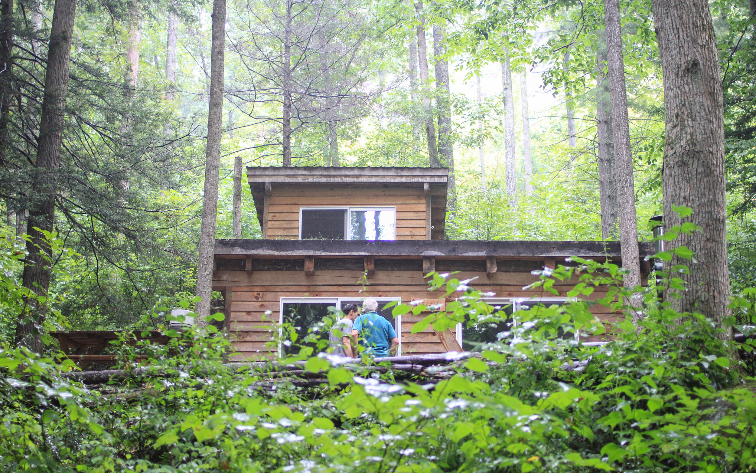 Two people standing on a wooden porch in front of a two-story, rustic wooden cabin surrounded by dense green forest.