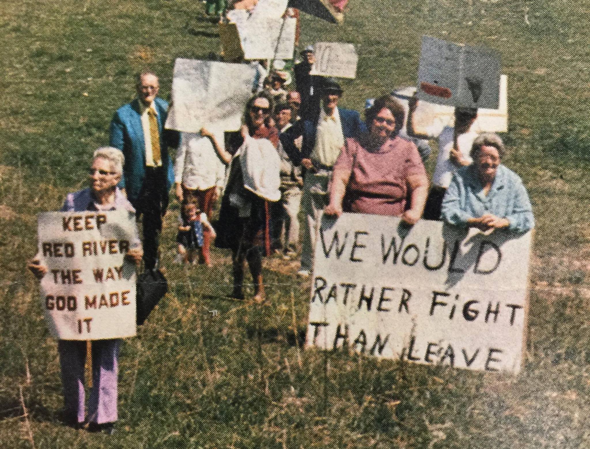A group of people at an outdoor protest or demonstration, holding handmade signs. The signs have messages such as "Keep Red River the way God made it" and "We would rather fight than leave." The group includes men, women, and children standing on a grassy area, some smiling and engaging with the camera.