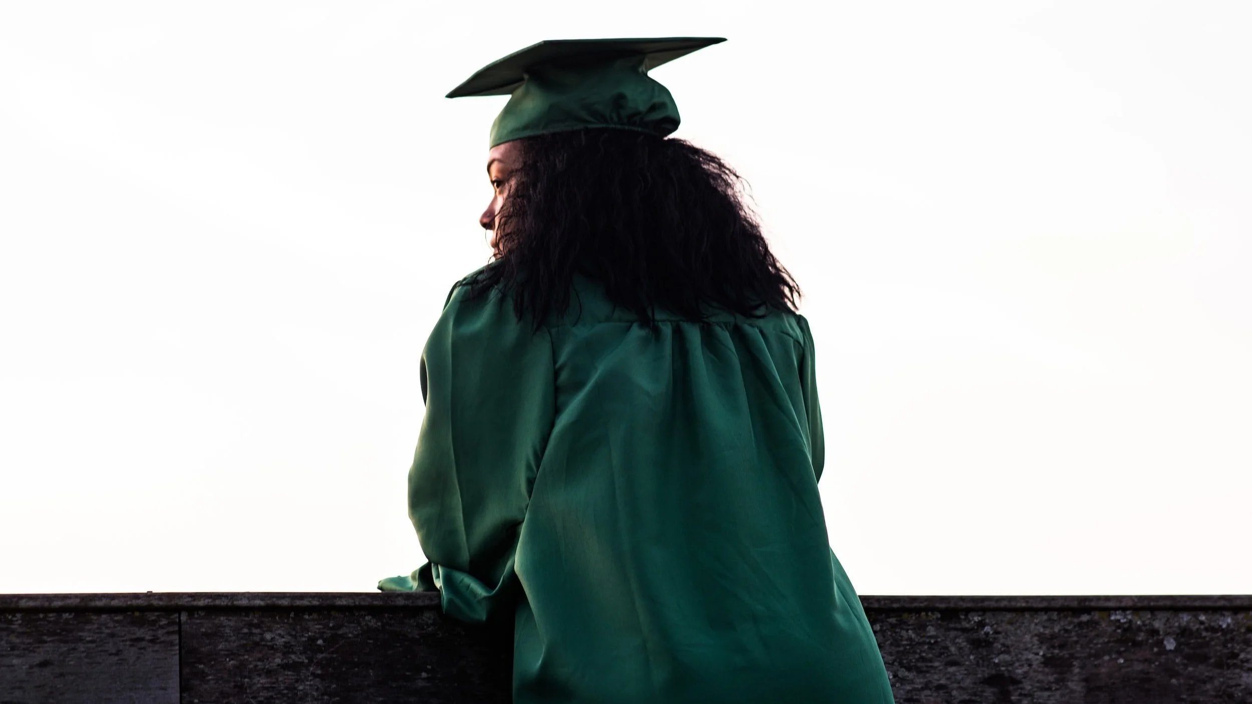 A woman in green graduation gown and cap standing outdoors on a ledge, facing sideways against a bright sky.