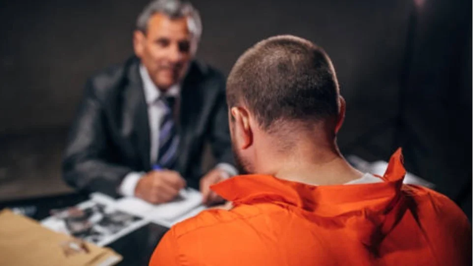 A man in an orange jumpsuit facing an older man in a suit during an interview or interrogation.