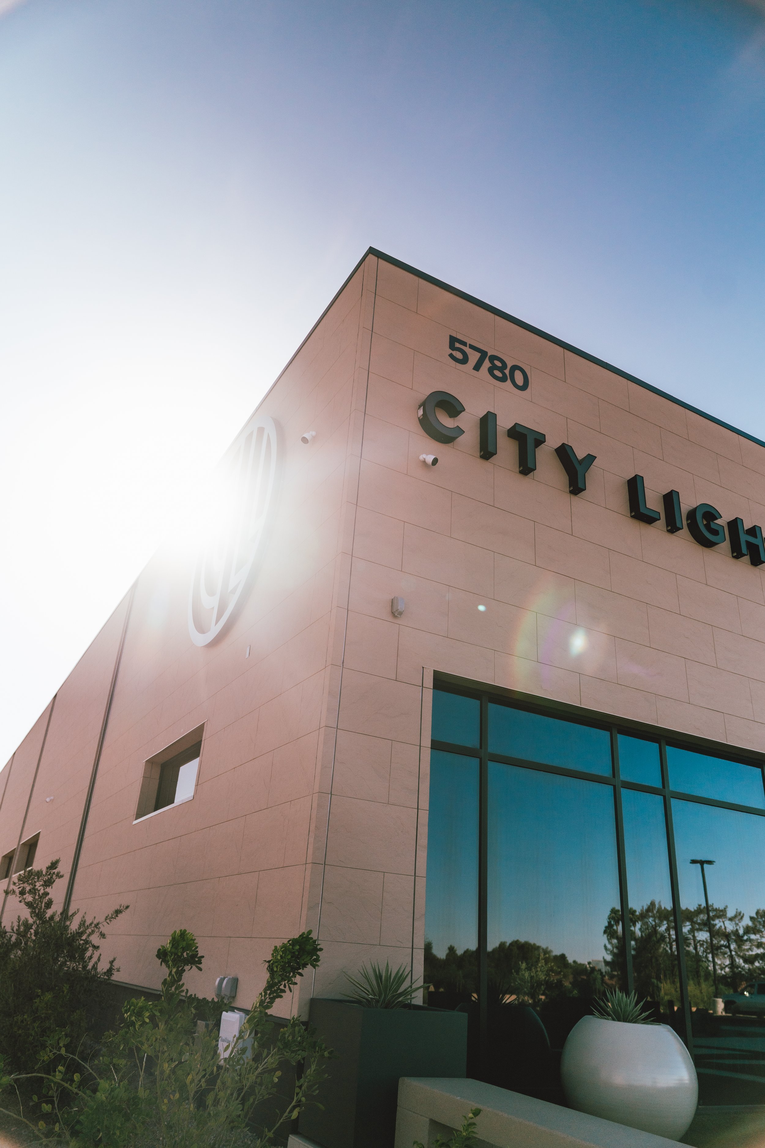 Exterior of a building with a sign that reads 'City Light' and the number 5780. Bright sunlight causing lens flare, with some plants and large windows reflecting the sky and trees.