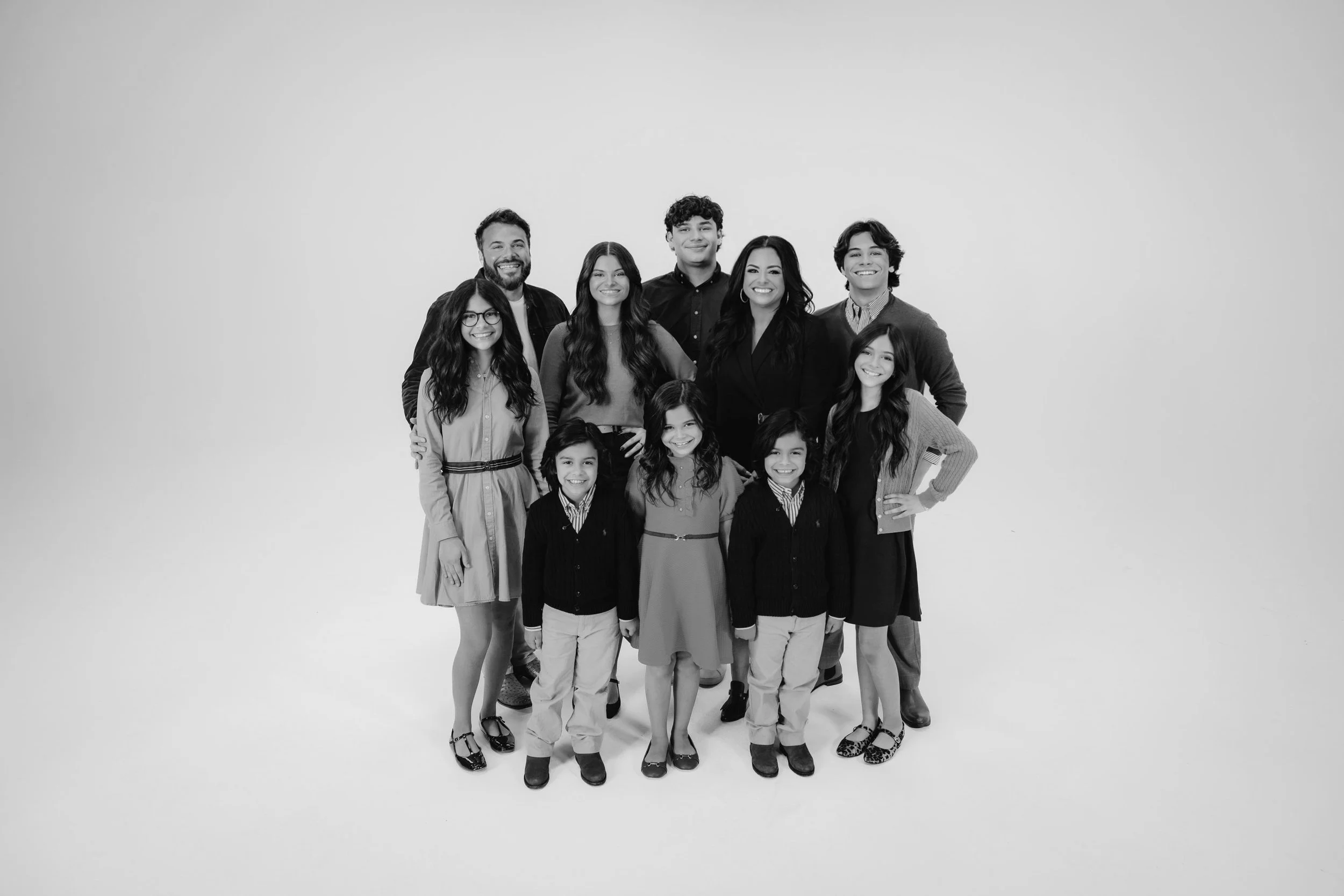 Black and white photo of a diverse group of eleven people, including adults and children, standing together and smiling in a studio with a plain background.