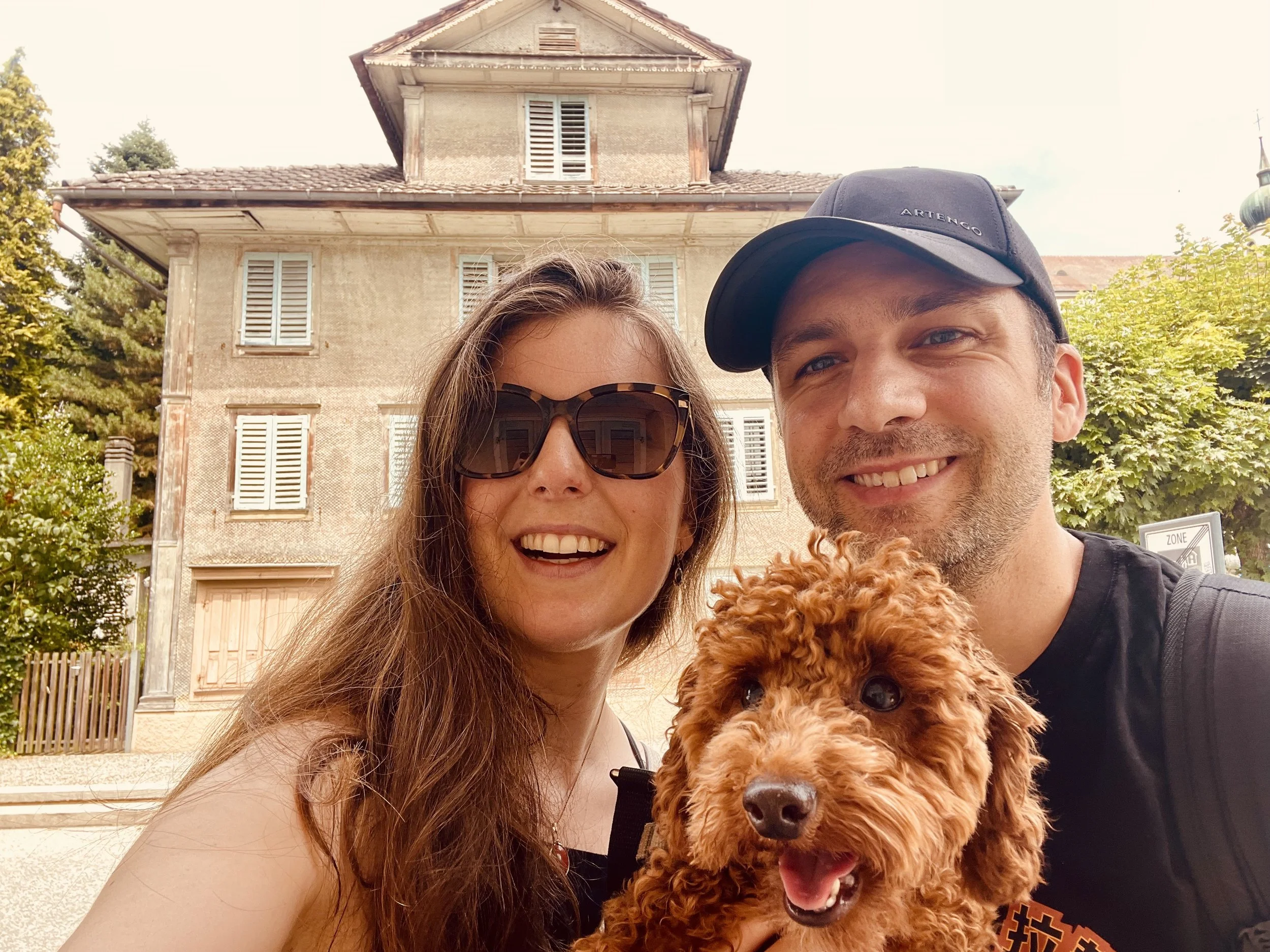 Simone McIntosh, Jean-Philippe Mc Clish and their auburn toy poodle smile in a selfie. They are standing in front of a historical house, seen in the distance. They are in a small Swiss town called Eschenbach.