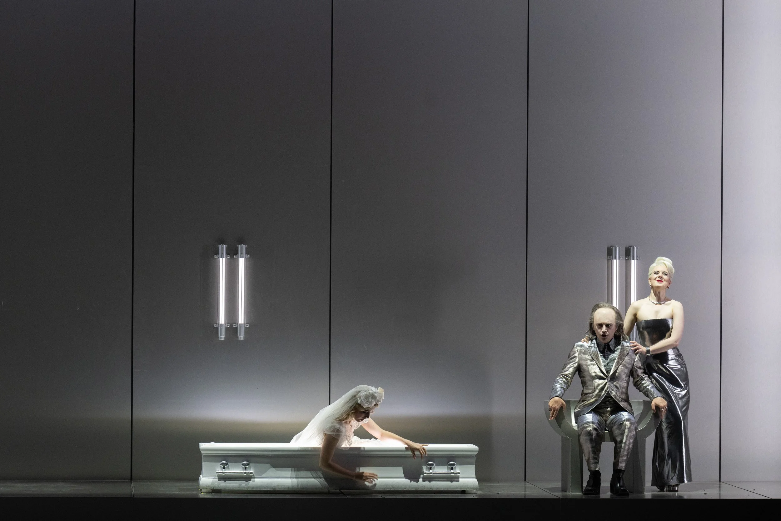 Simone McIntosh, Mirco Palazzi and Miriam Kutrowatz are in a scene in front of a grey, futuristic background. Mirco is sitting in a metal chair, Simone sits on the arm of the chair holding him proudly. Miriam arises from a white coffin.