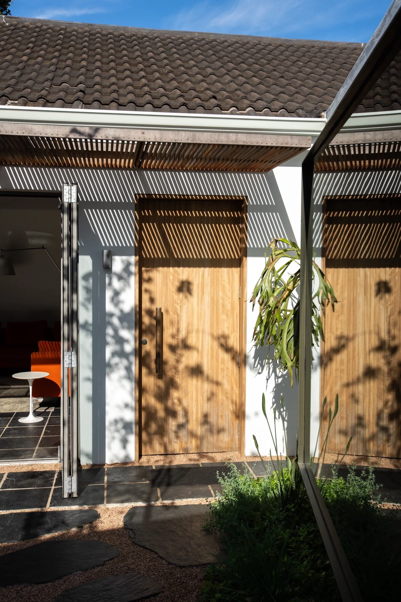 Exterior view of a house featuring a wooden door, a sliding glass door leading to a living room, a slanted roof with tiles, and shadows cast by outdoor plants and a pergola.